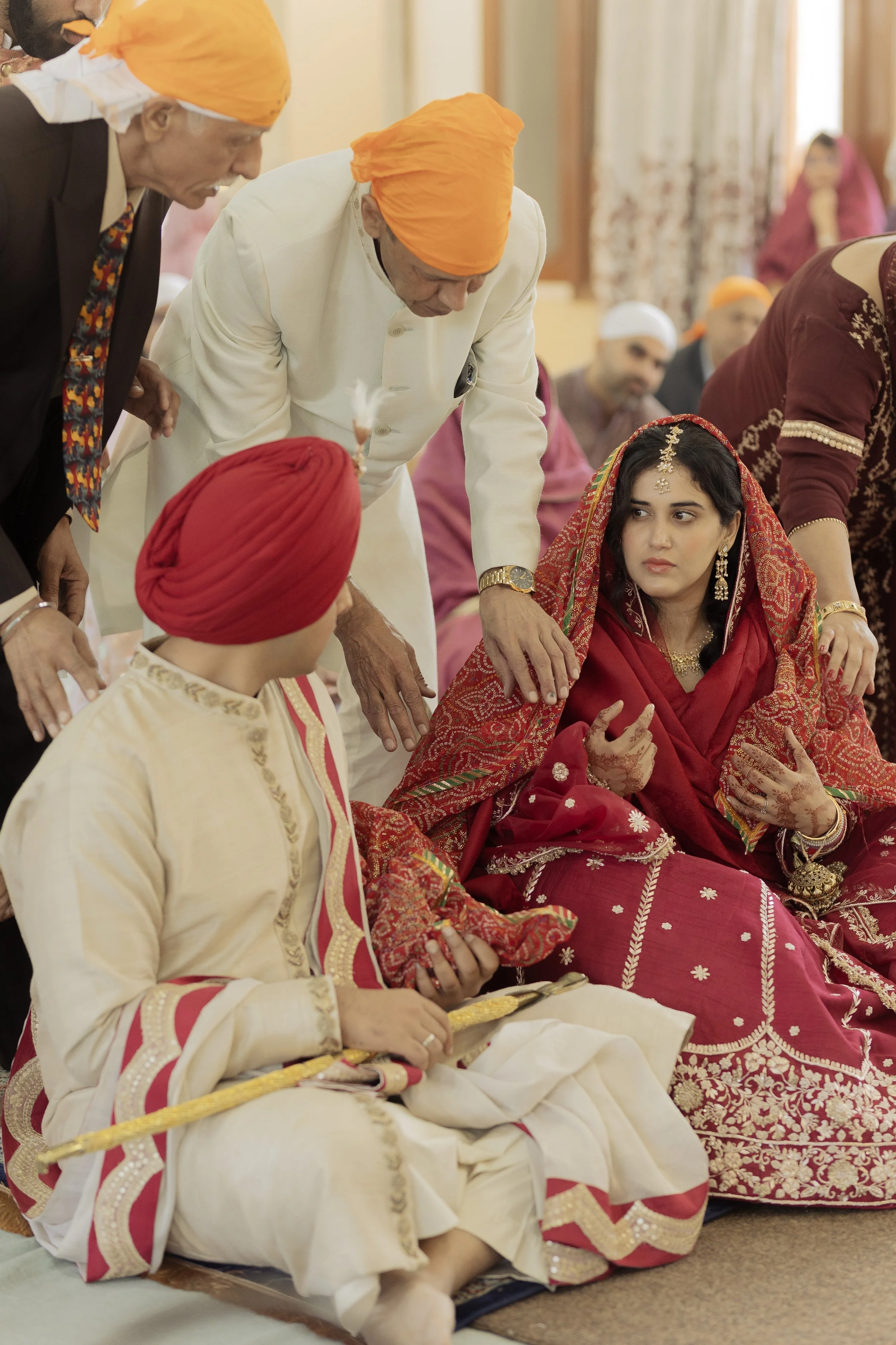 Indian wedding ceremony with a woman in a red saree and multiple men in traditional attire, some wearing orange turbans, gathered indoors.