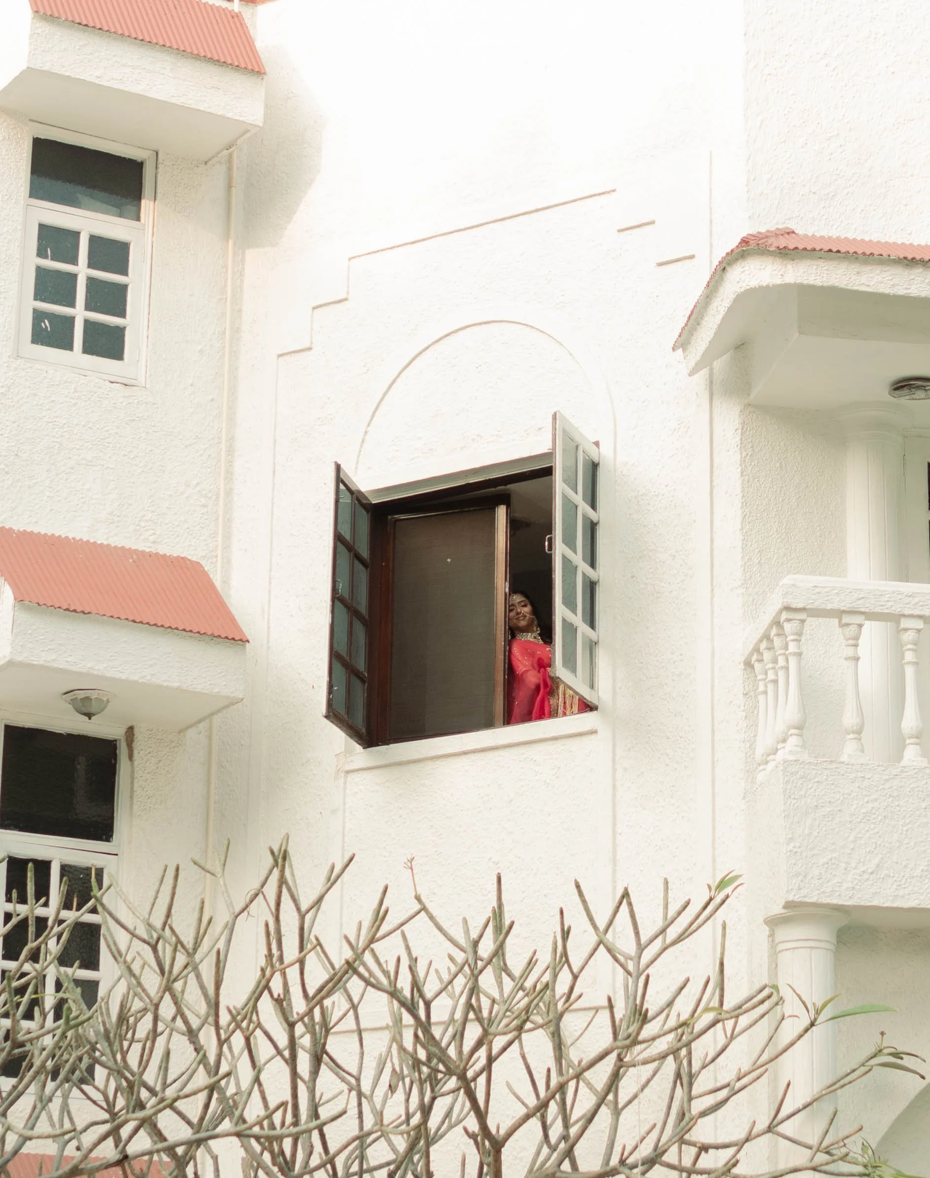 A woman in a pink dress standing at an open window of a white building, looking outside.