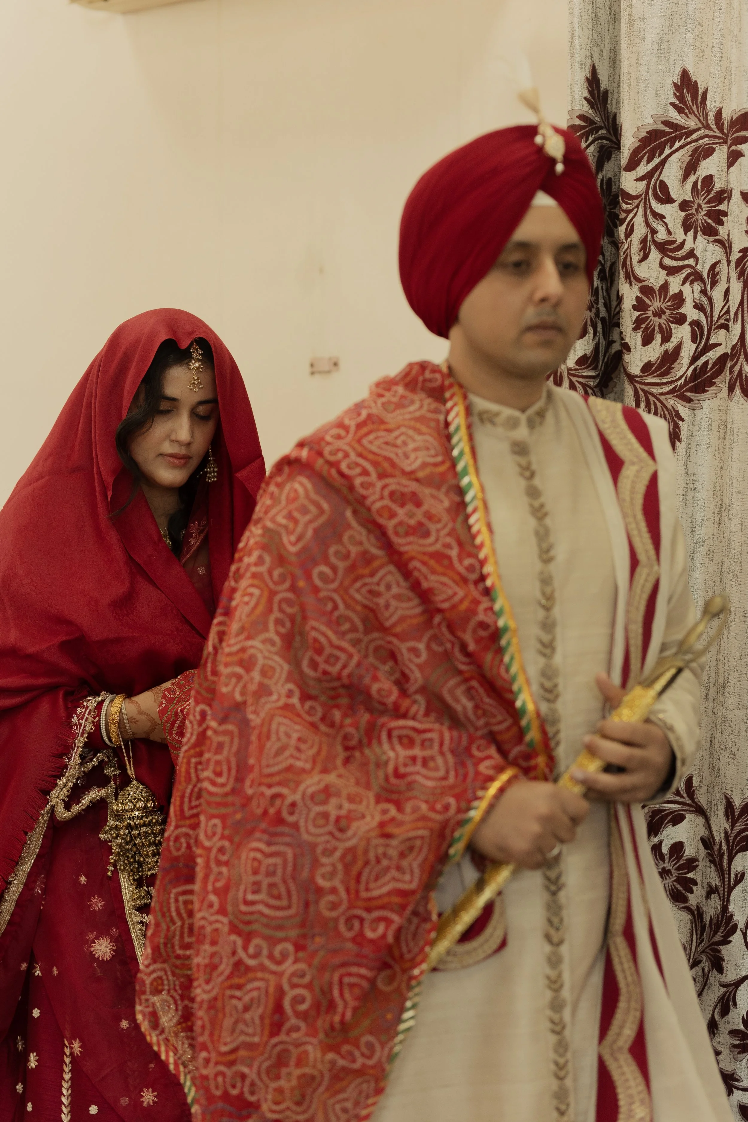 A man and woman dressed in traditional Indian wedding attire, standing indoors; the woman in a red saree with gold jewelry and a head covering, and the man in a cream-colored outfit with a red turban, holding a staff, with a floral curtain in the bac