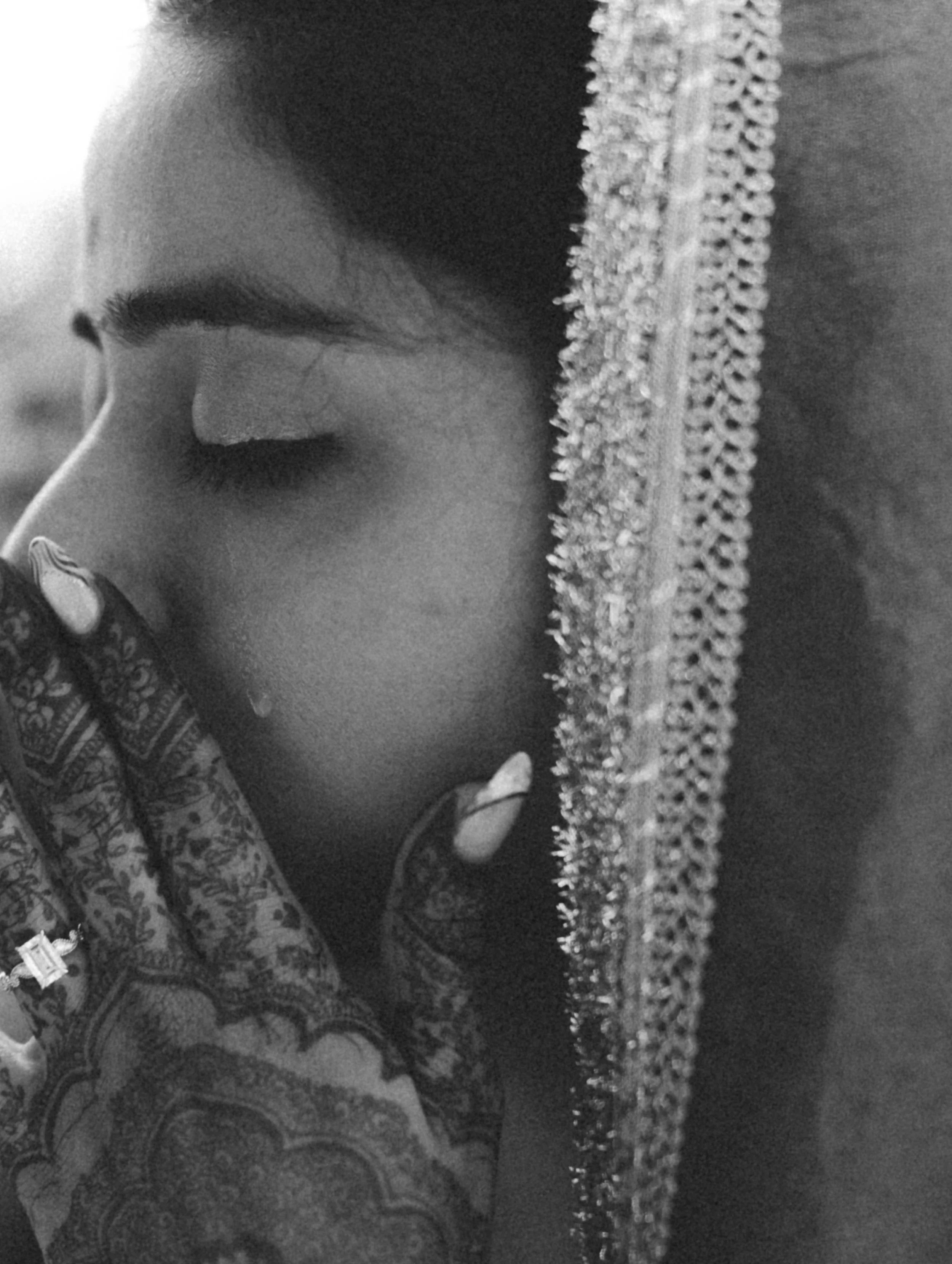 Close-up black-and-white photo of a woman with her eyes closed, wearing a veil and intricately patterned henna on her hand.