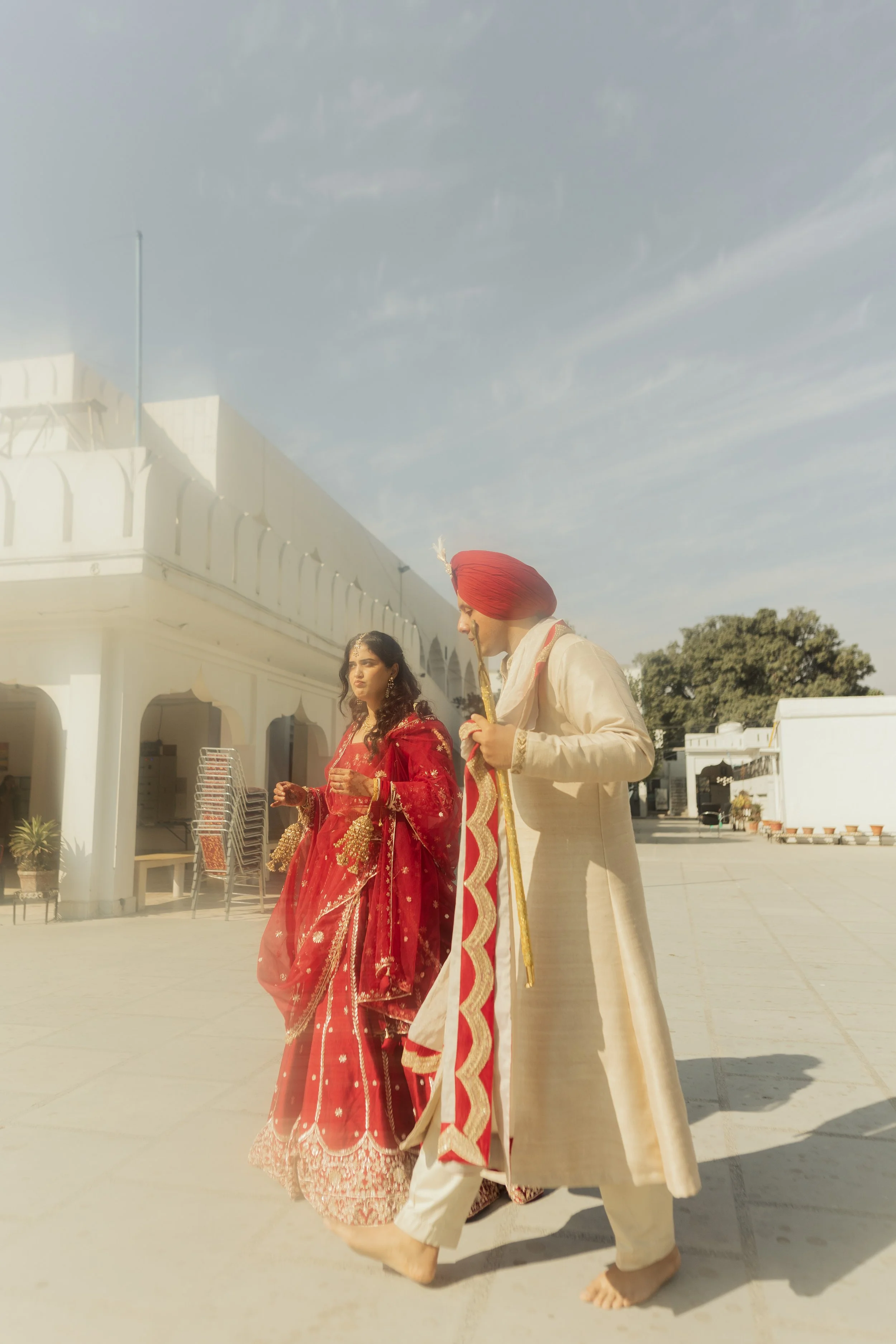A man and a woman in traditional Indian attire standing outdoors. The woman is dressed in a red embellished saree, and the man is in a cream-colored sherwani with a red turban. They are holding hands and appear to be participating in a wedding ceremo