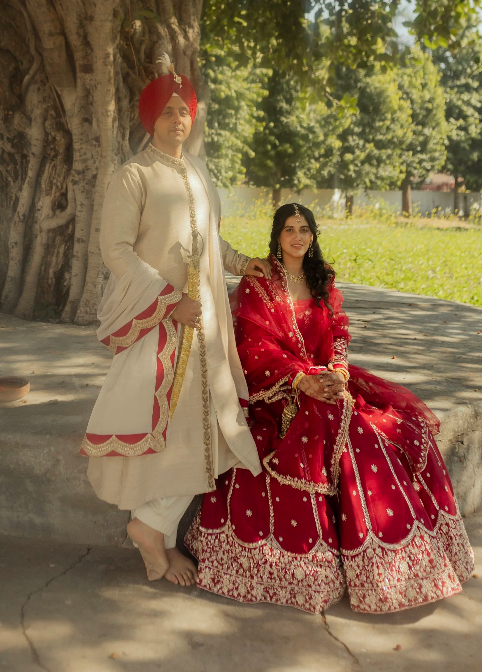 A man dressed in traditional Indian wedding attire standing next to a woman in a vibrant red wedding dress, sitting on a stone ledge outdoors under a large tree, with green grass and trees in the background.