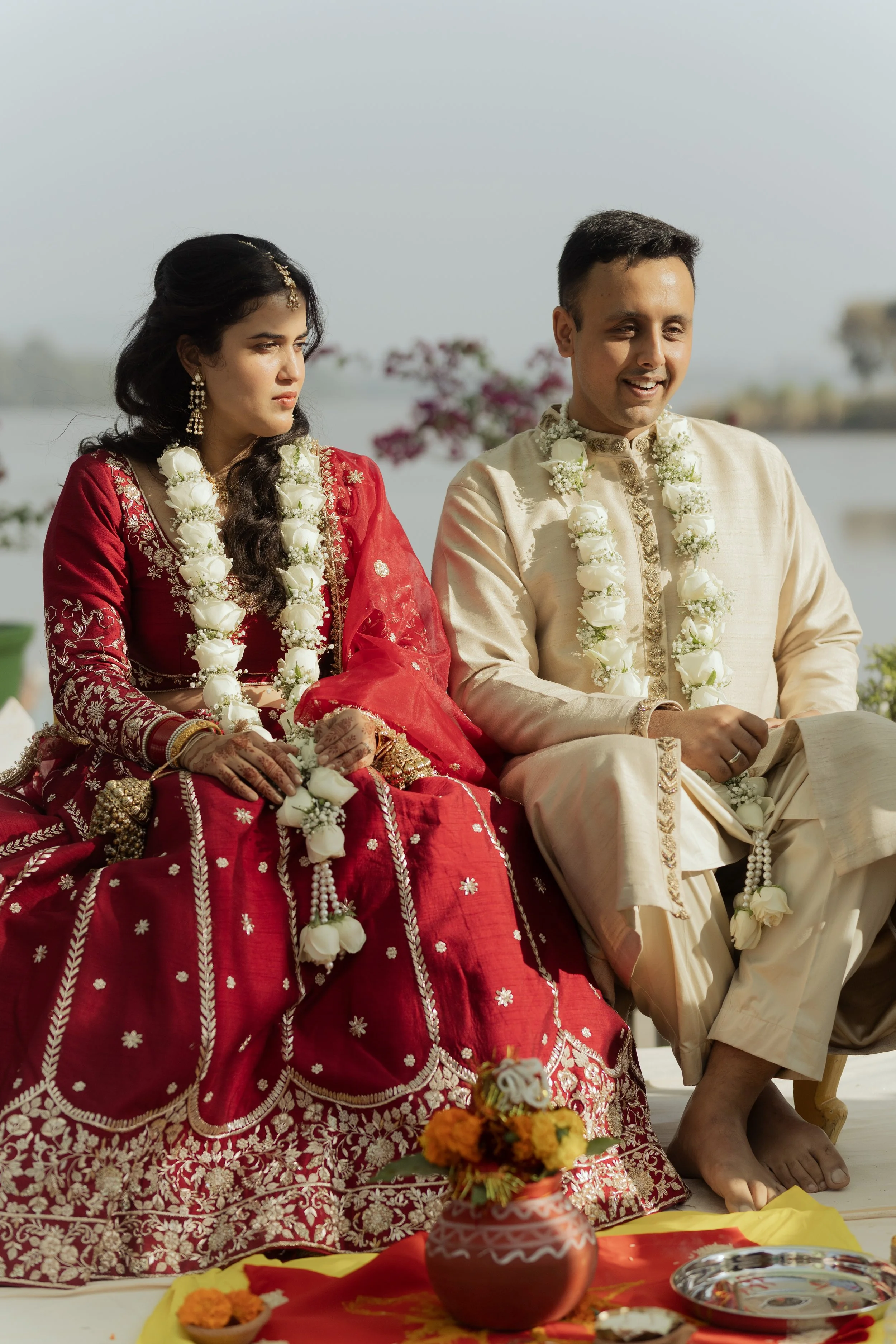 A South Asian bride and groom dressed in traditional wedding attire, sitting outdoors. The bride wears a red embroidered dress and jewelry, and the groom wears a cream-colored traditional outfit. Both are adorned with white flower garlands.