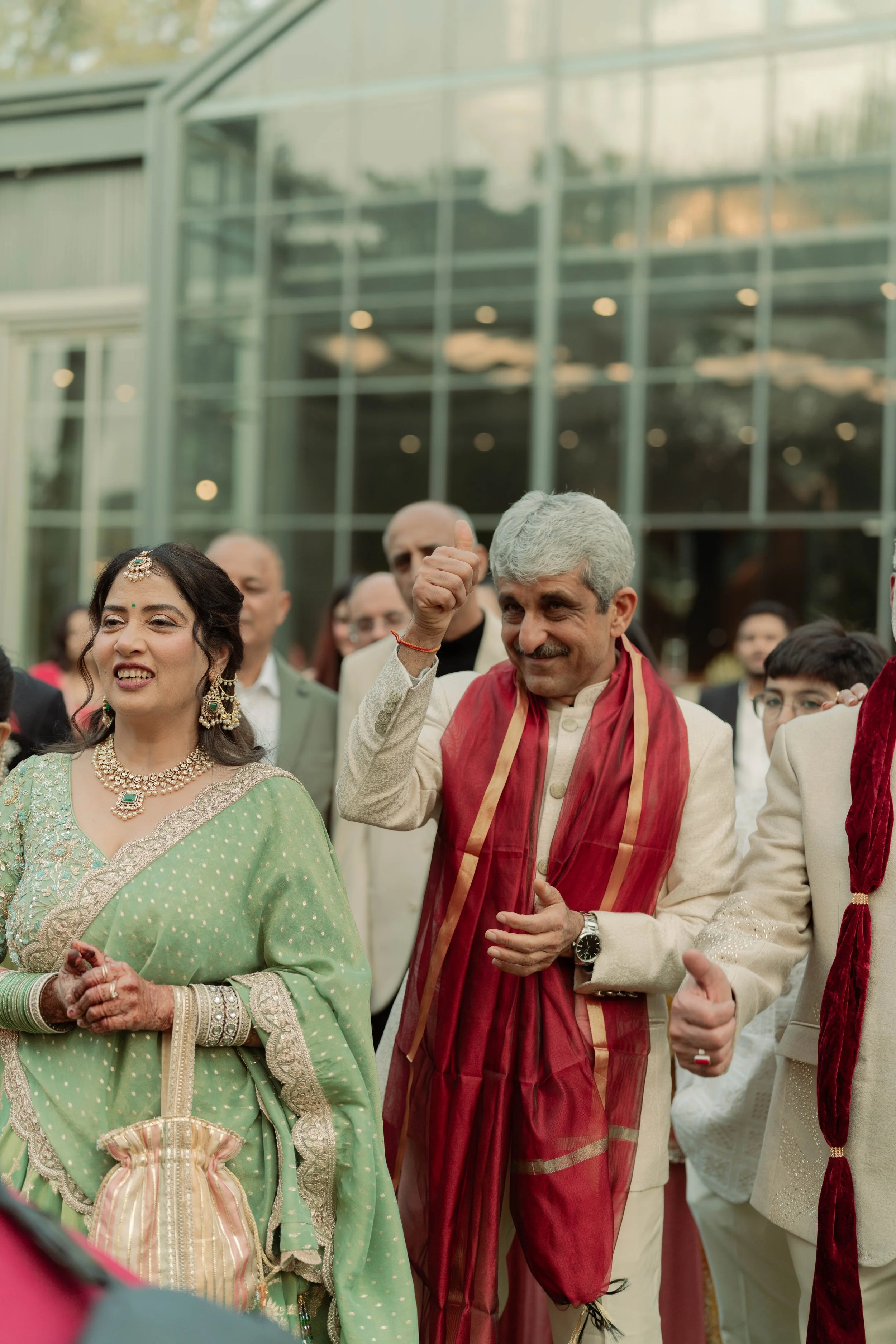 Indian man and woman in traditional attire attending a celebration outdoors, surrounded by other attendees, in front of a modern glass building.