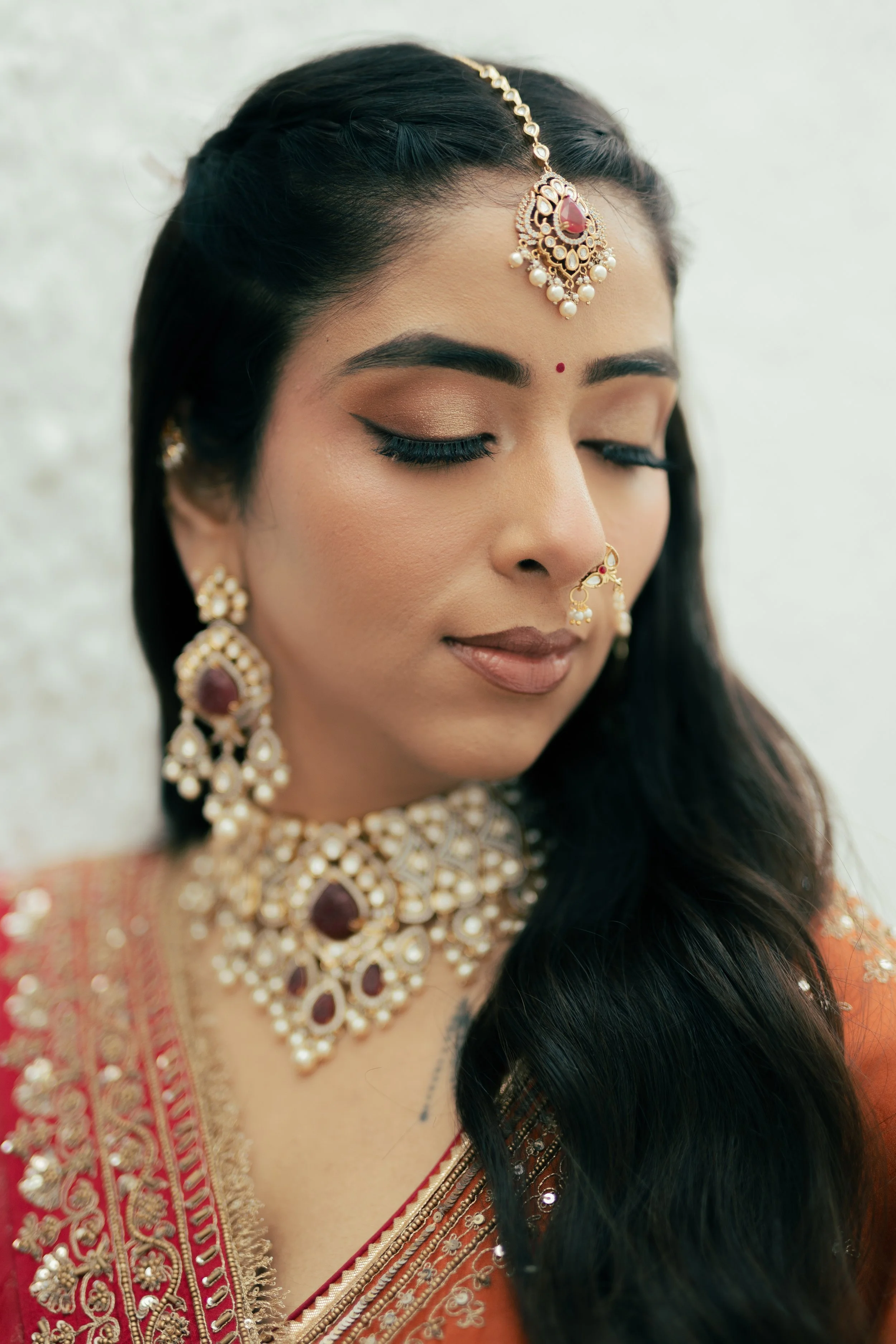 Close-up of a woman wearing traditional Indian jewelry, including a maang tikka, earrings, nose ring, and necklace, with her eyes closed and a serene expression, dressed in a richly embroidered orange and gold saree.