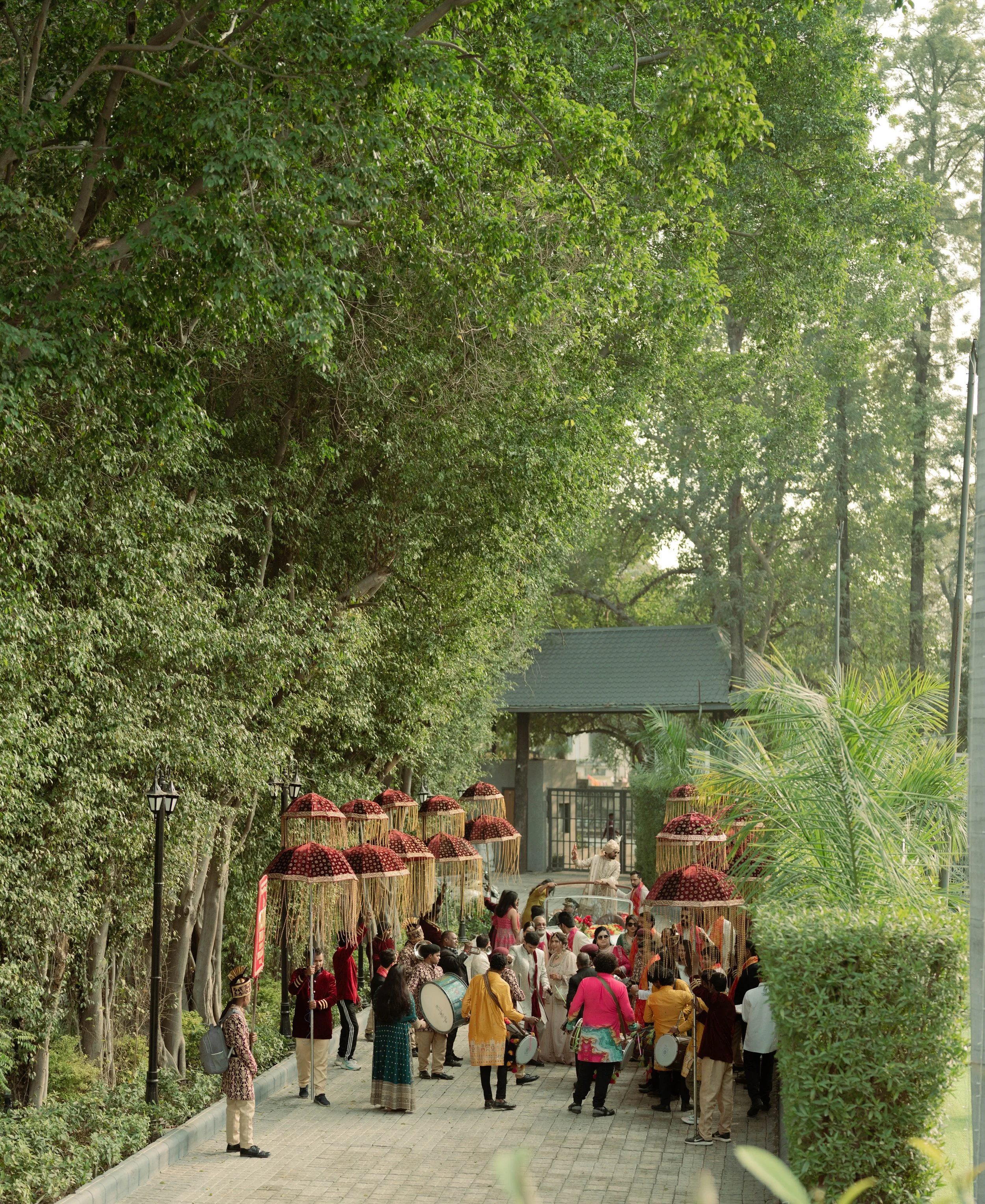 Group of people participating in a cultural or traditional celebration outdoors with umbrellas and drums, surrounded by green trees.