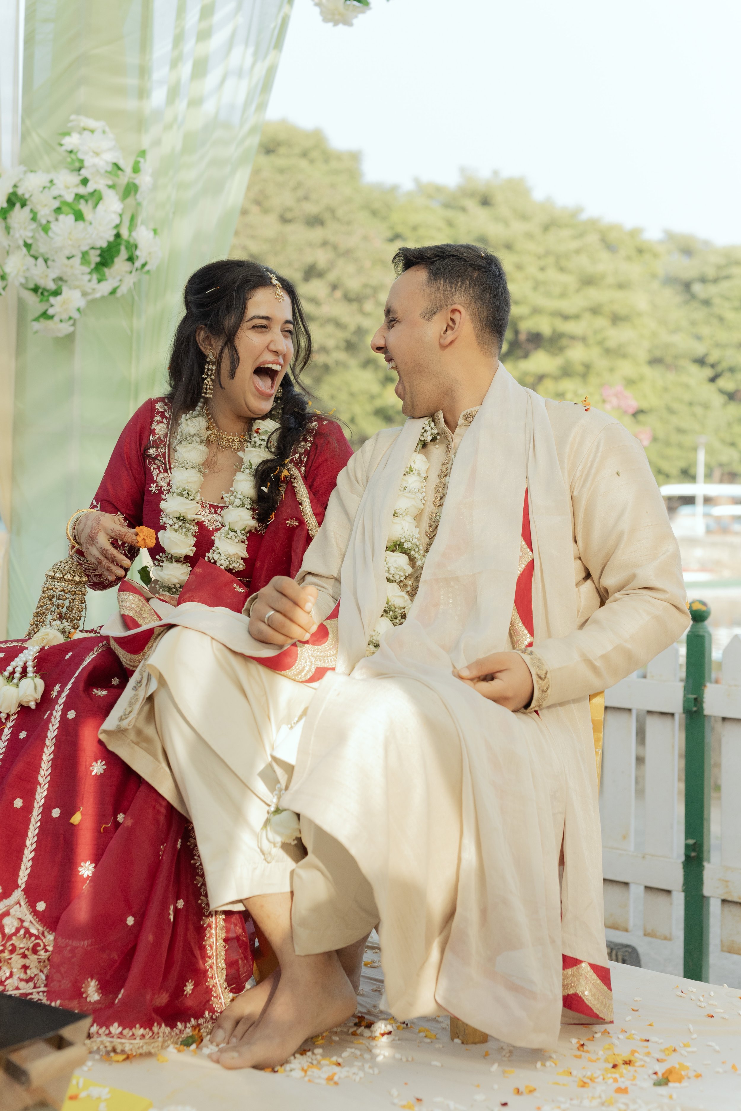 A couple participating in a traditional Indian wedding ceremony, sitting outdoors, joyfully smiling and laughing, dressed in traditional Indian attire with floral garlands around their necks.