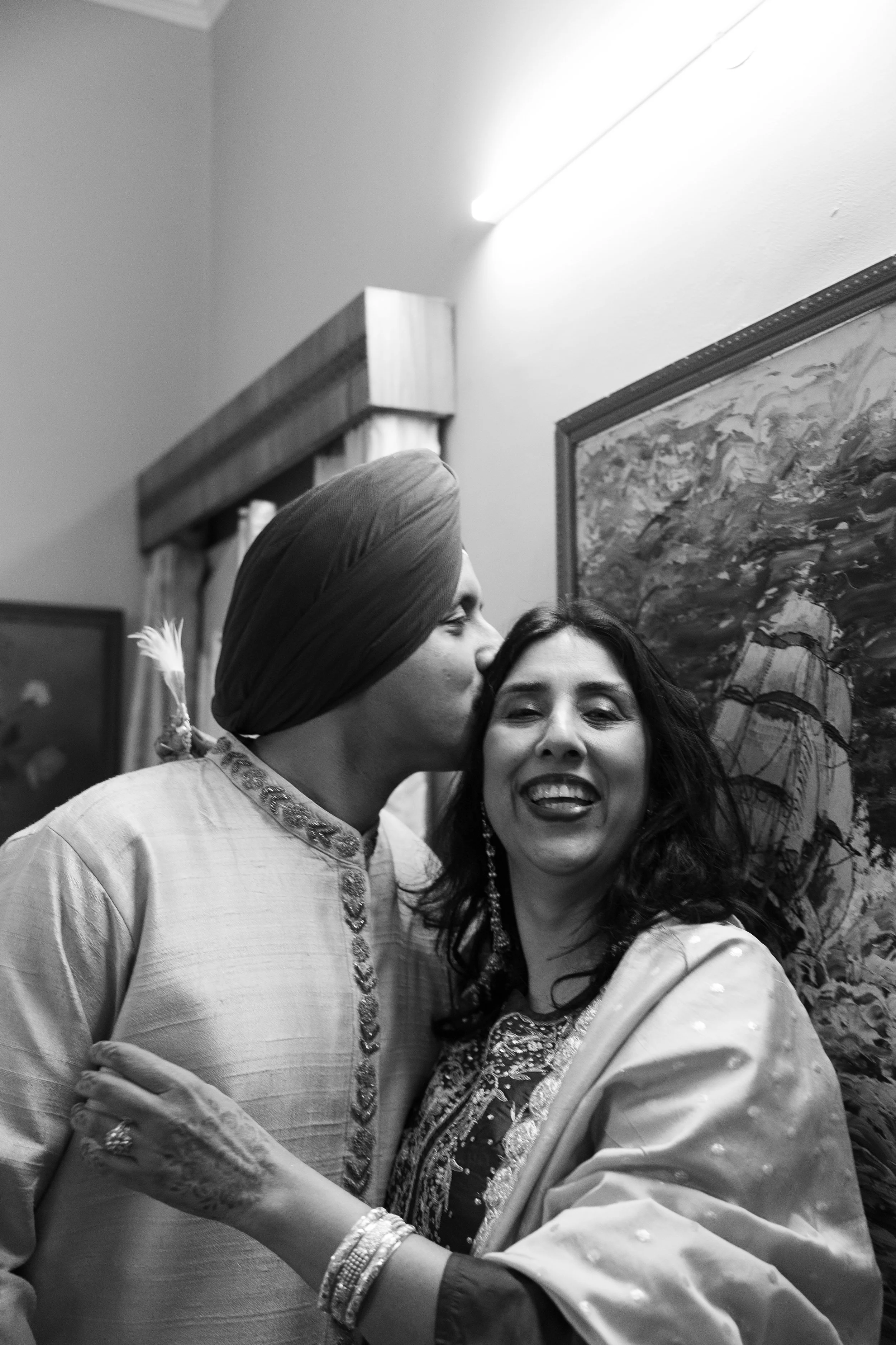 A man wearing a turban kisses a smiling woman on the cheek, both in traditional Indian attire, indoors with a painting on the wall behind them.