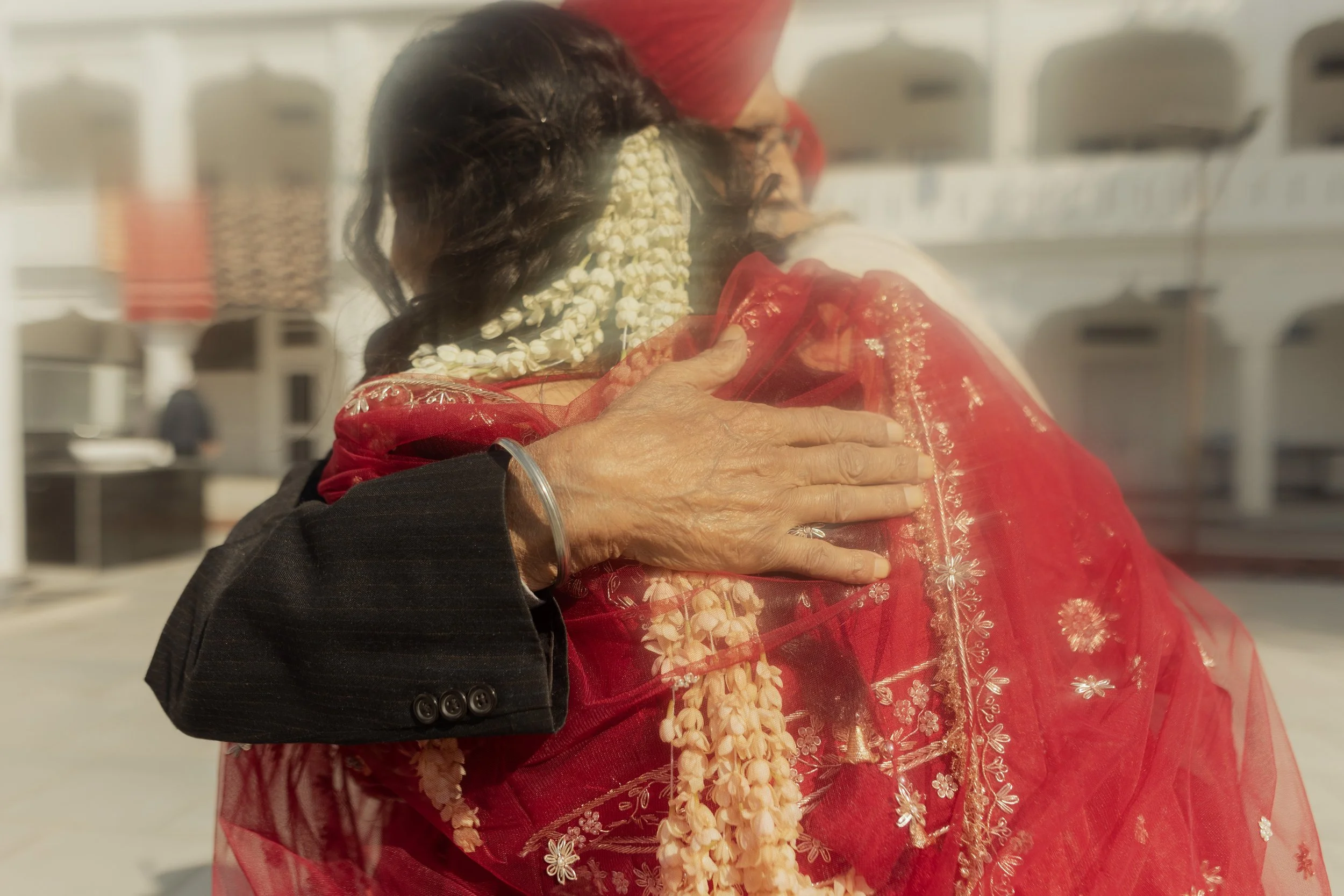 An elderly woman in traditional red Indian attire is hugging a man in a dark suit. The woman is wearing a saree with gold embroidery and flowers, and has a garland around her neck. The man’s hand, with a silver bracelet, is resting on her back.