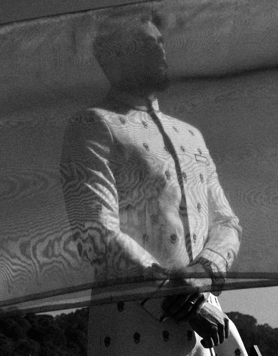 Black and white photo of a man wearing a chef's coat, standing outdoors with trees in the background, holding a phone or device in his hands.