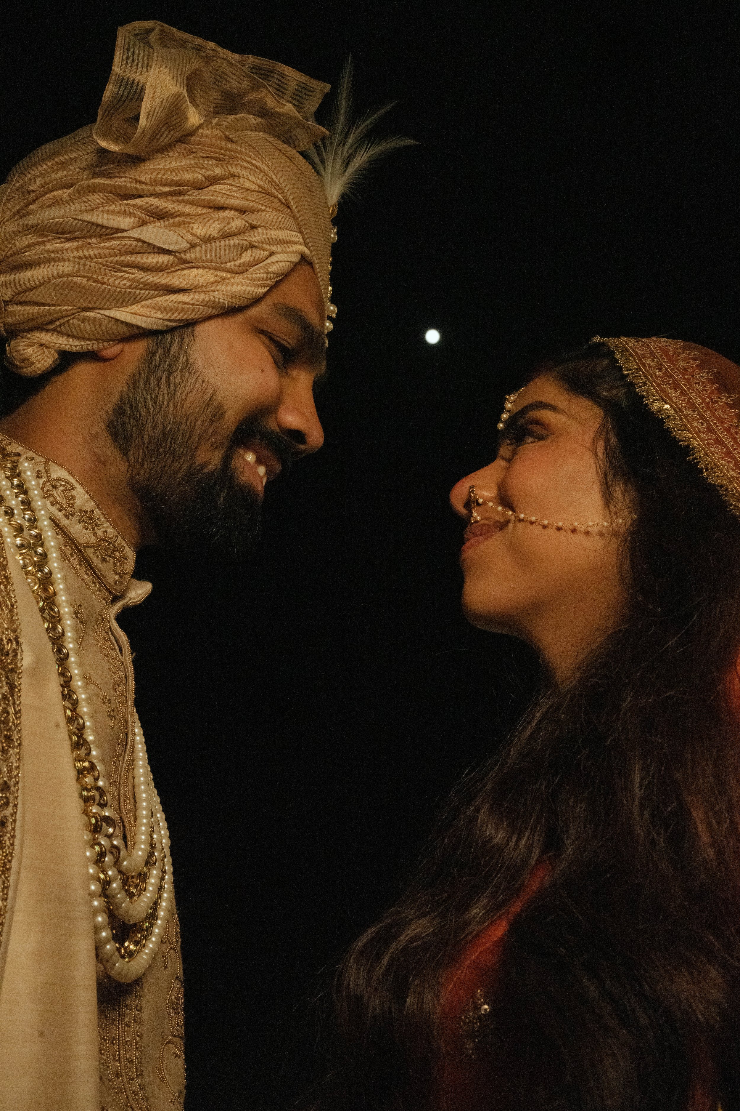 A couple dressed in traditional Indian wedding attire, sharing an intimate moment under the night sky with the moon visible.
