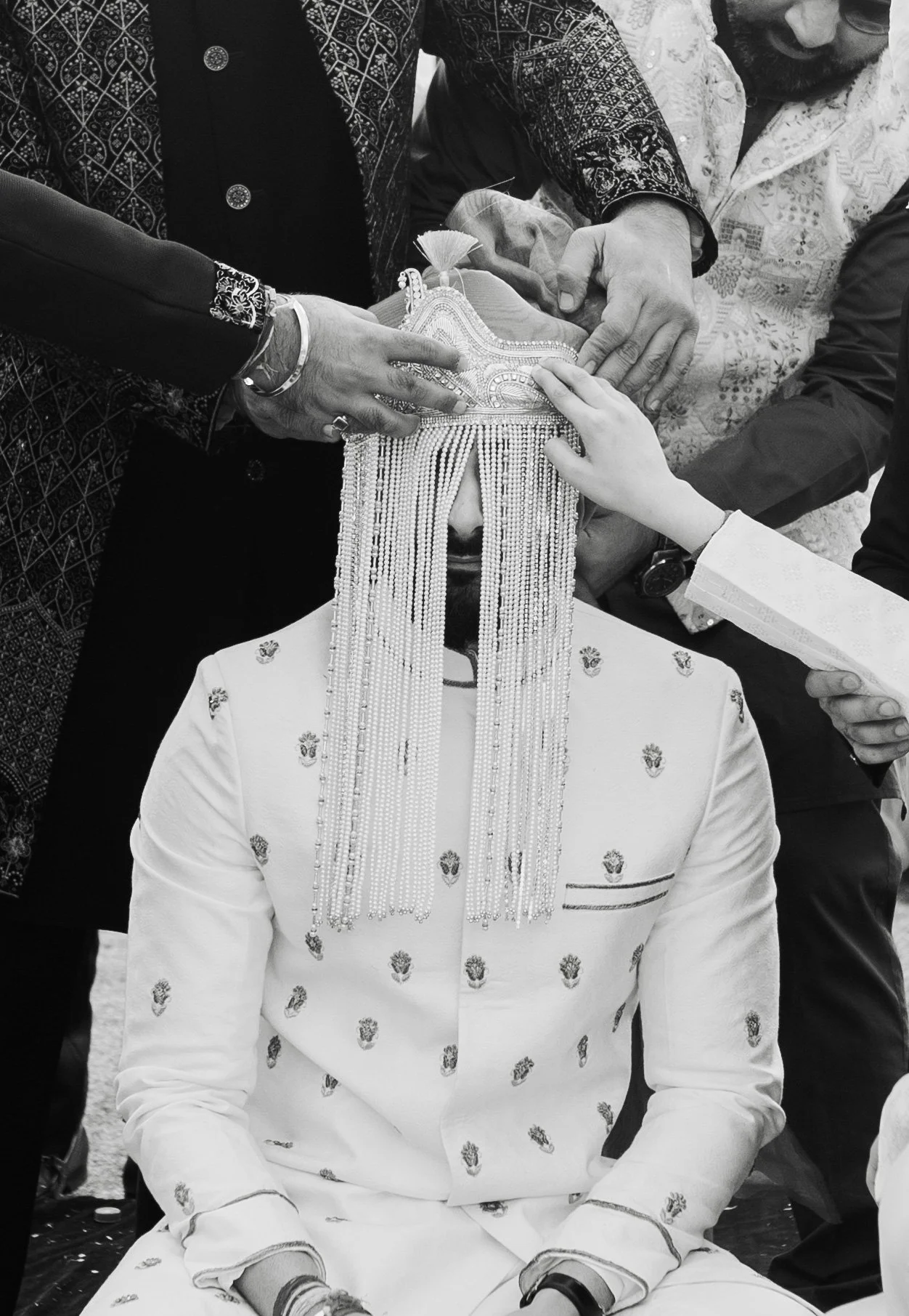 Man dressed in traditional Indian wedding attire participating in a wedding ritual with multiple hands placing a crown or headpiece on his head.