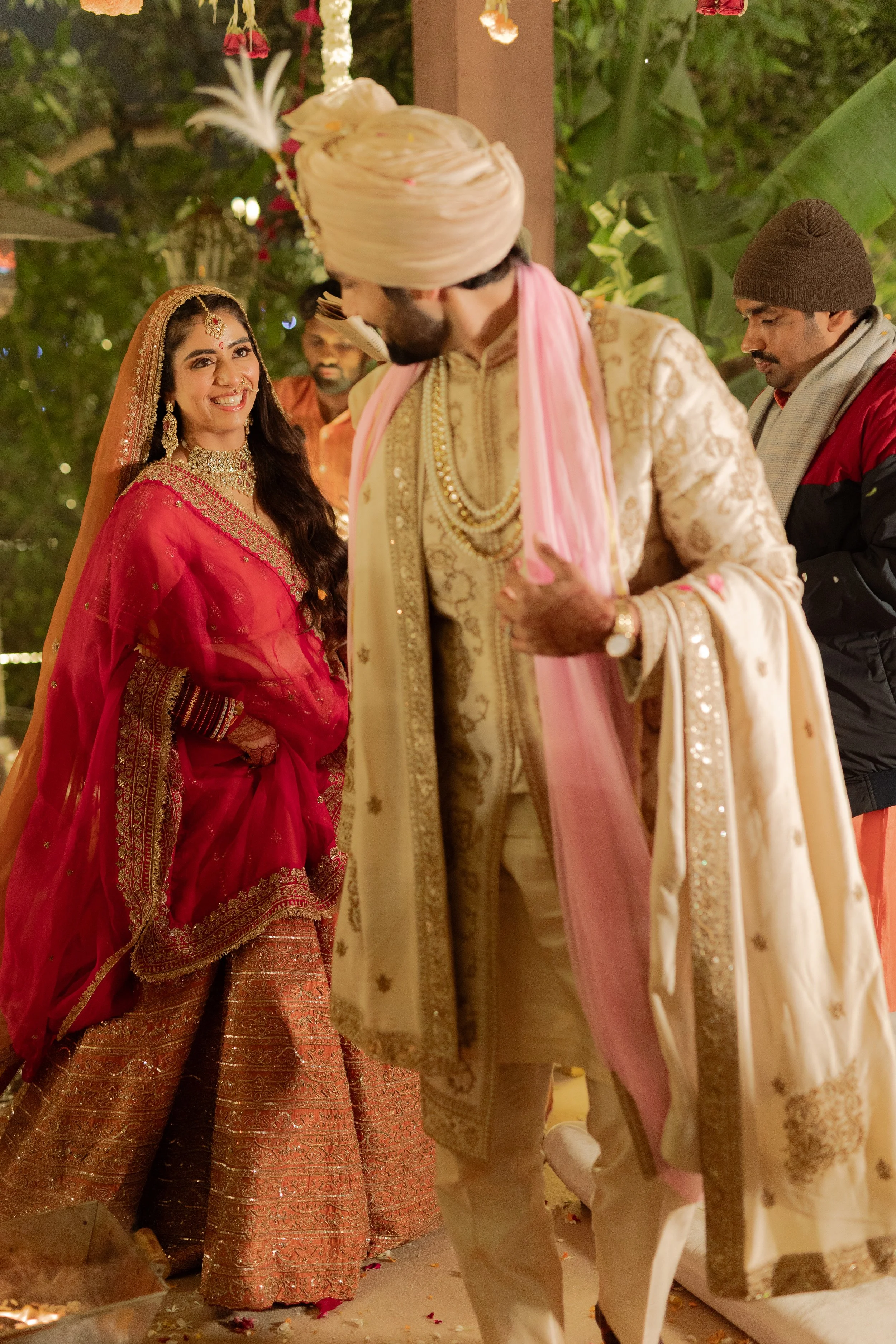 A bride dressed in a red and gold traditional wedding outfit and jewelry smiling at a groom dressed in an ornate cream and gold sherwani with a pink dupatta, during an Indian wedding ceremony.