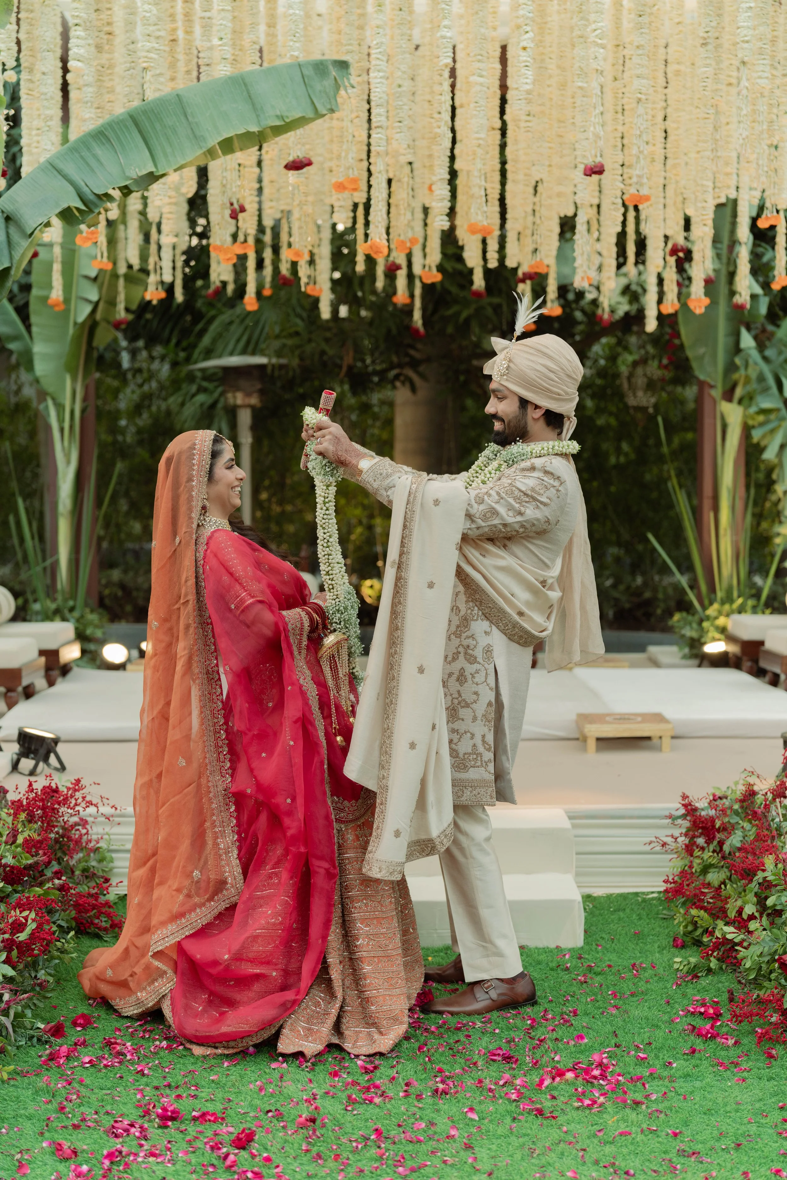 A bride and groom in traditional Indian wedding attire exchanging garlands during a wedding ceremony outdoors, with a decorated floral backdrop and greenery.