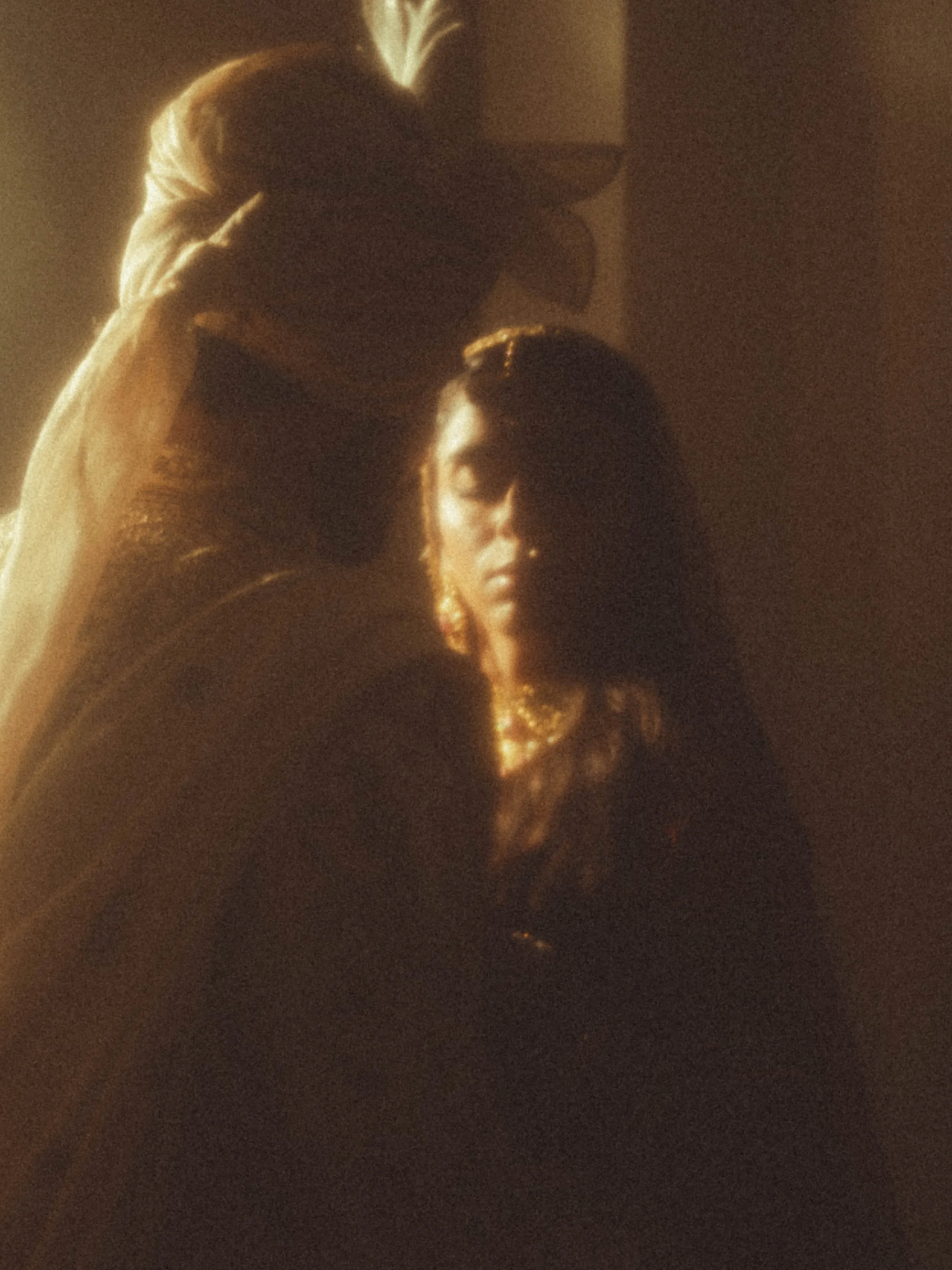 A woman with long dark hair and jewelry sitting with her eyes closed as a person places a cloth over her head in a dimly lit room.