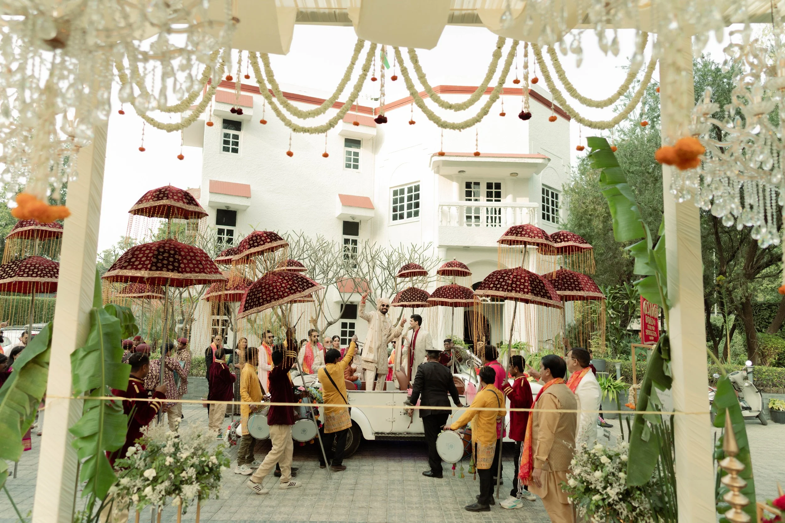 A group of people celebrates outdoors with traditional attire, holding umbrellas, near a white building with balconies, surrounded by greenery and decorated with flowers and hanging ornaments.