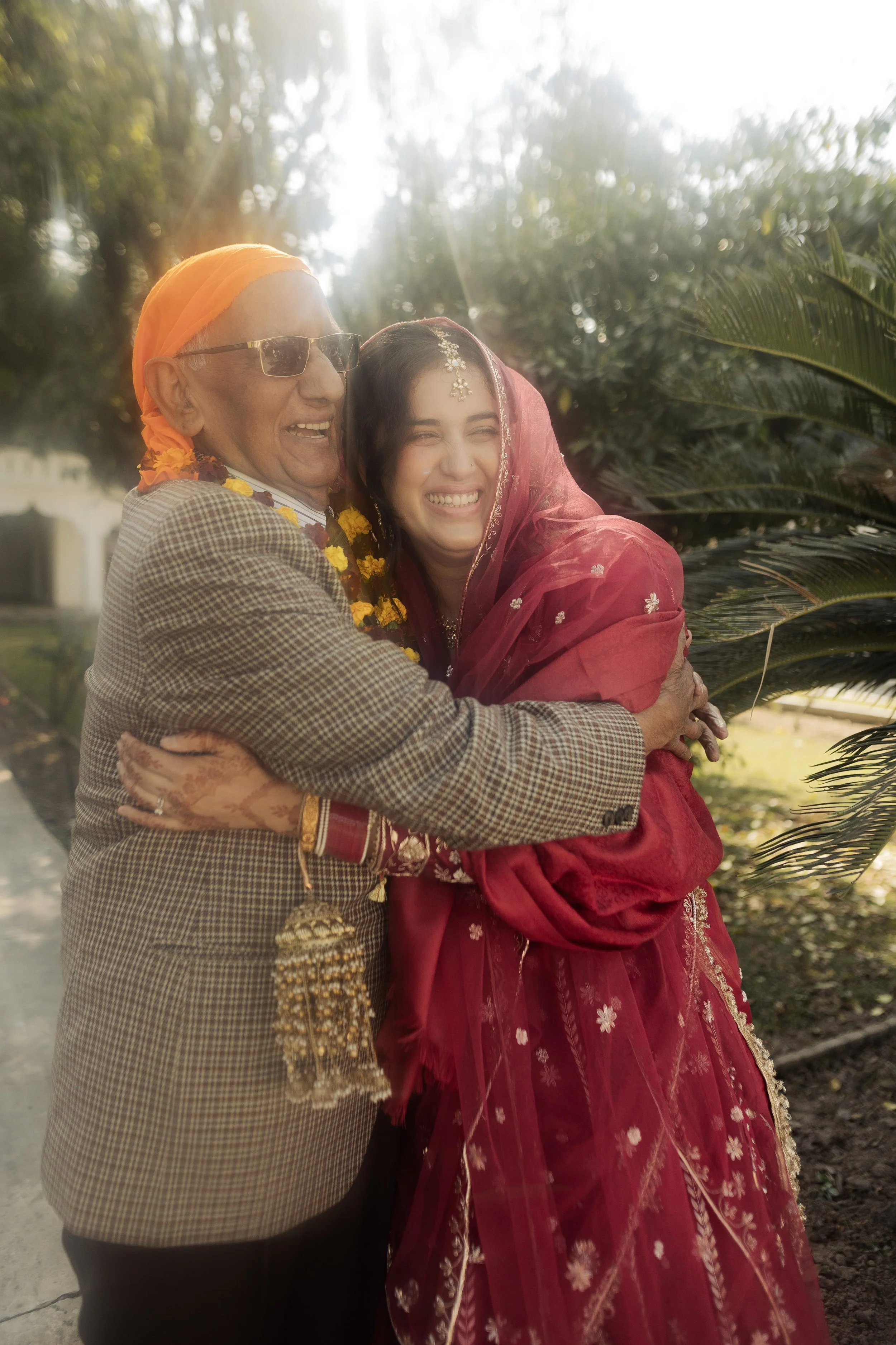 An elderly man and a young woman are hugging and smiling outdoors in bright sunlight, surrounded by greenery. The man is wearing sunglasses, a printed suit jacket, and a headscarf, while the woman is dressed in a red traditional outfit with jewelry a