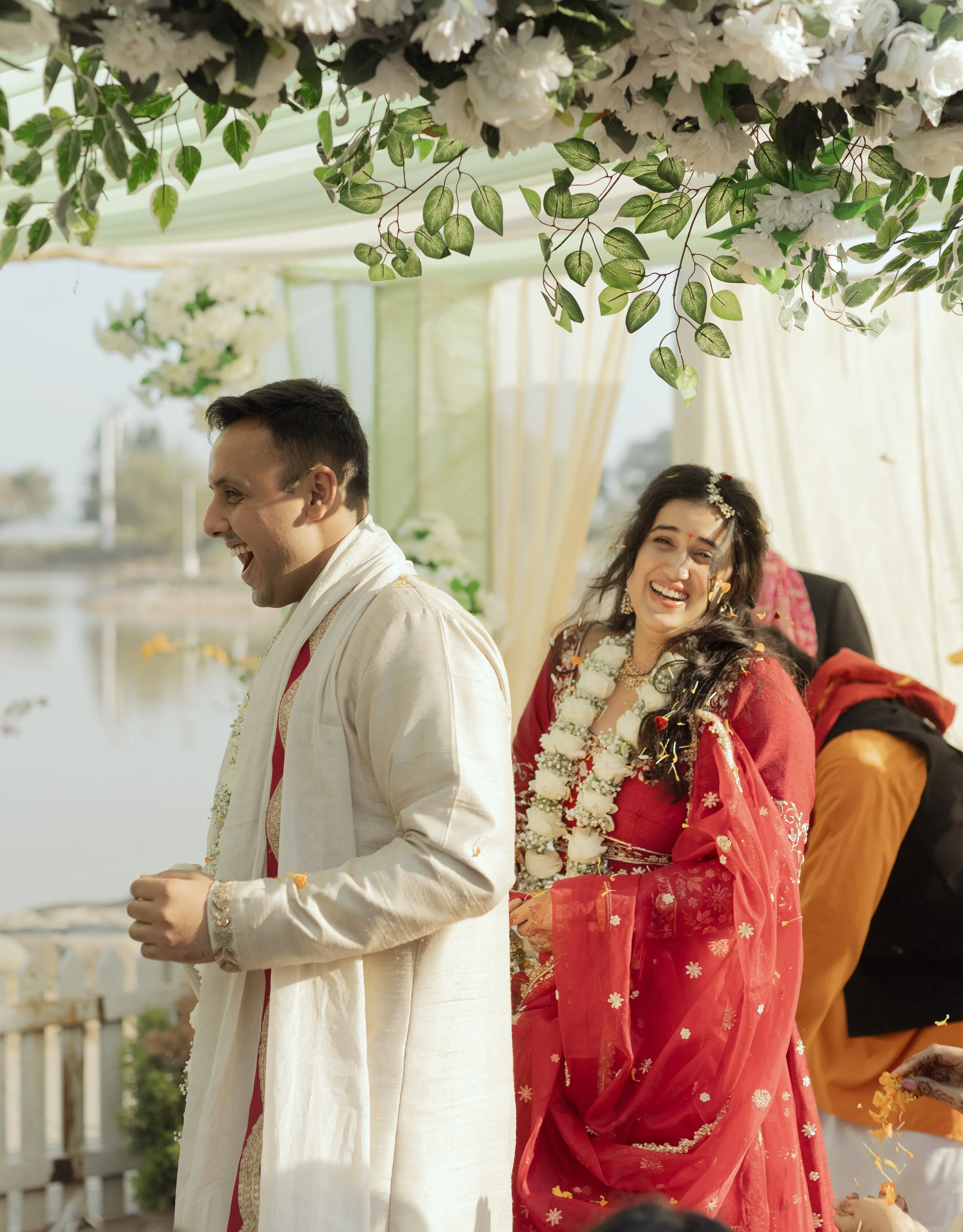 A couple at a wedding celebration, dressed in traditional Indian attire, smiling and celebrating under a decorated canopy near a body of water.