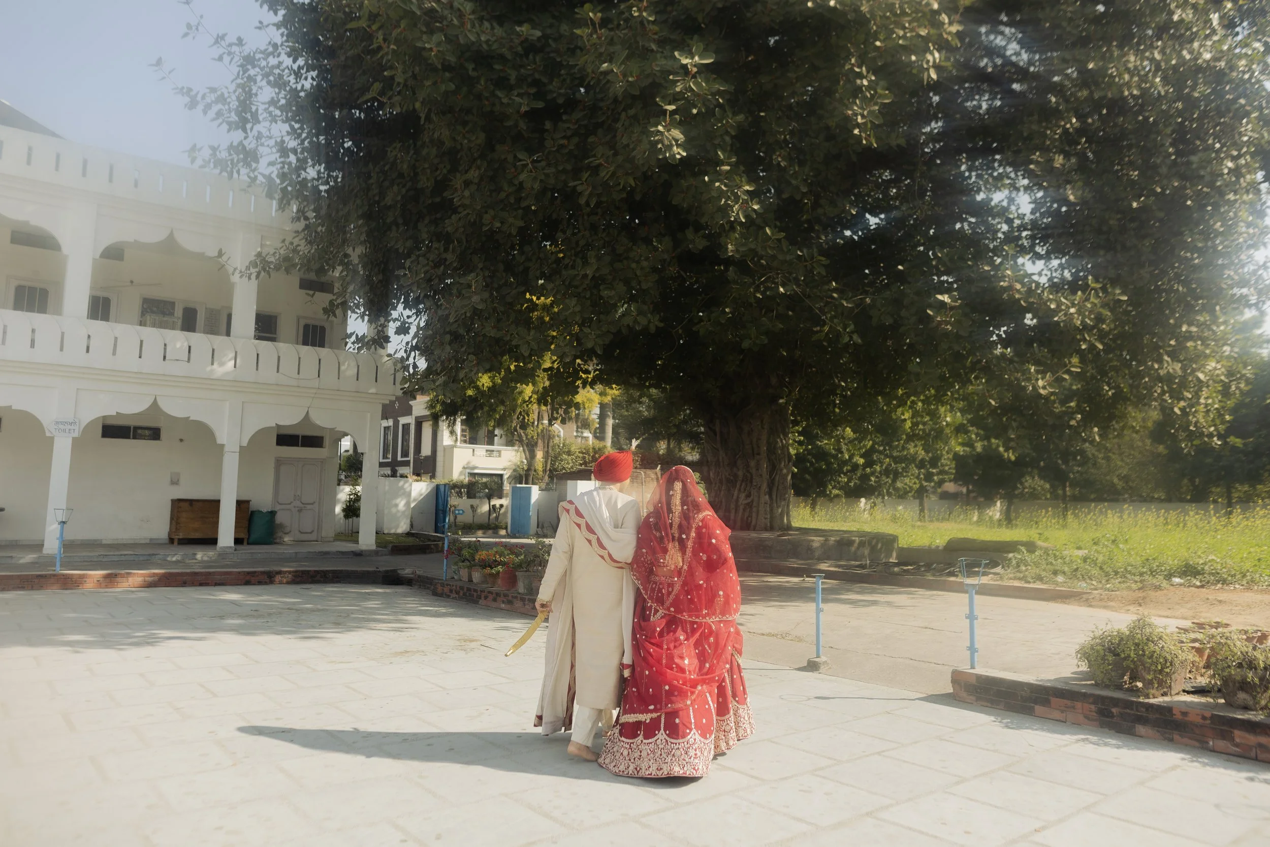 A couple dressed in traditional Indian wedding attire walking away from the camera, holding hands, under a large tree in a courtyard.