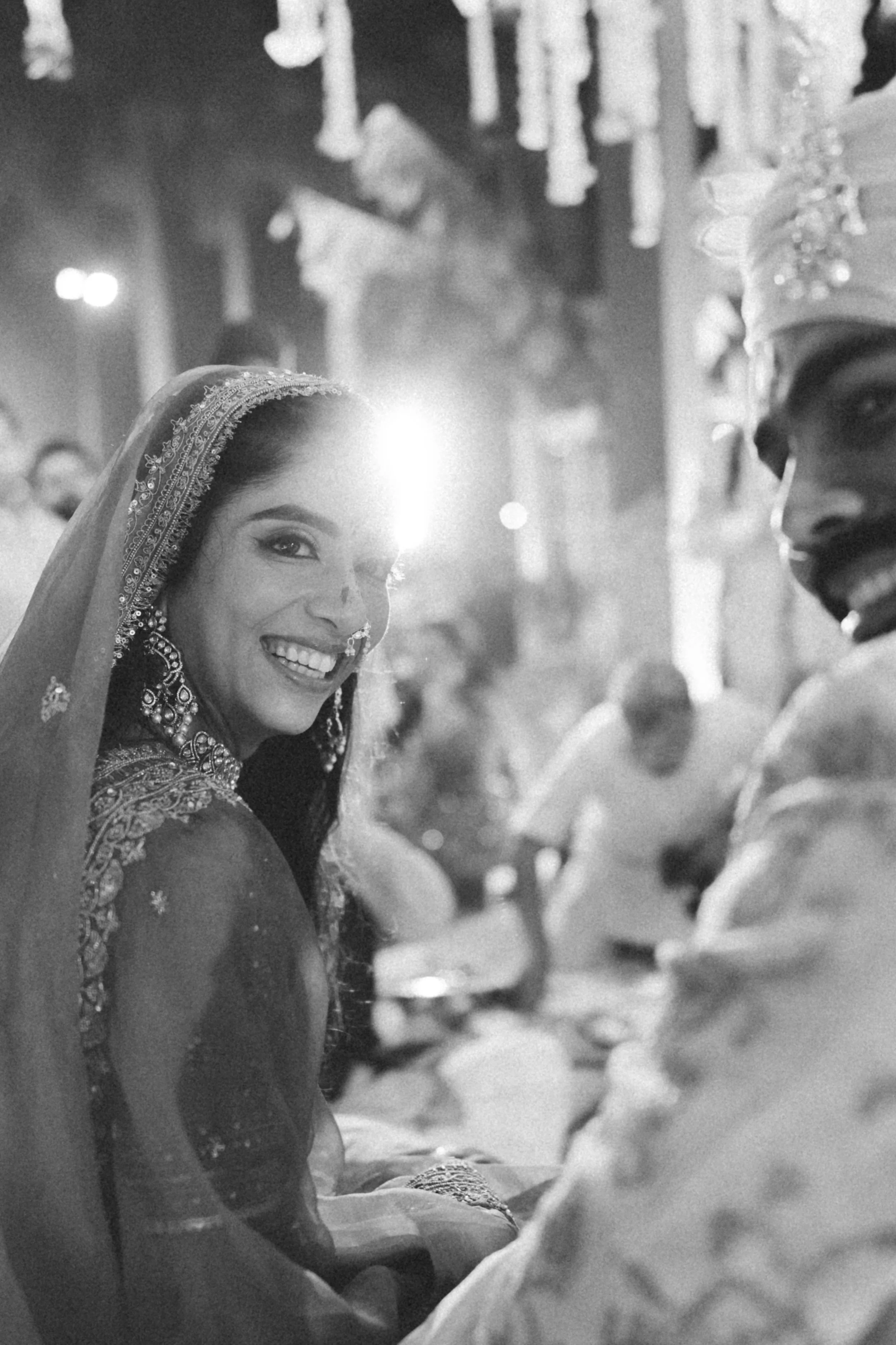 A smiling bride with traditional Indian jewelry and attire at a wedding, sitting with a groom in ceremonial clothing, surrounded by guests in an elaborately decorated venue.