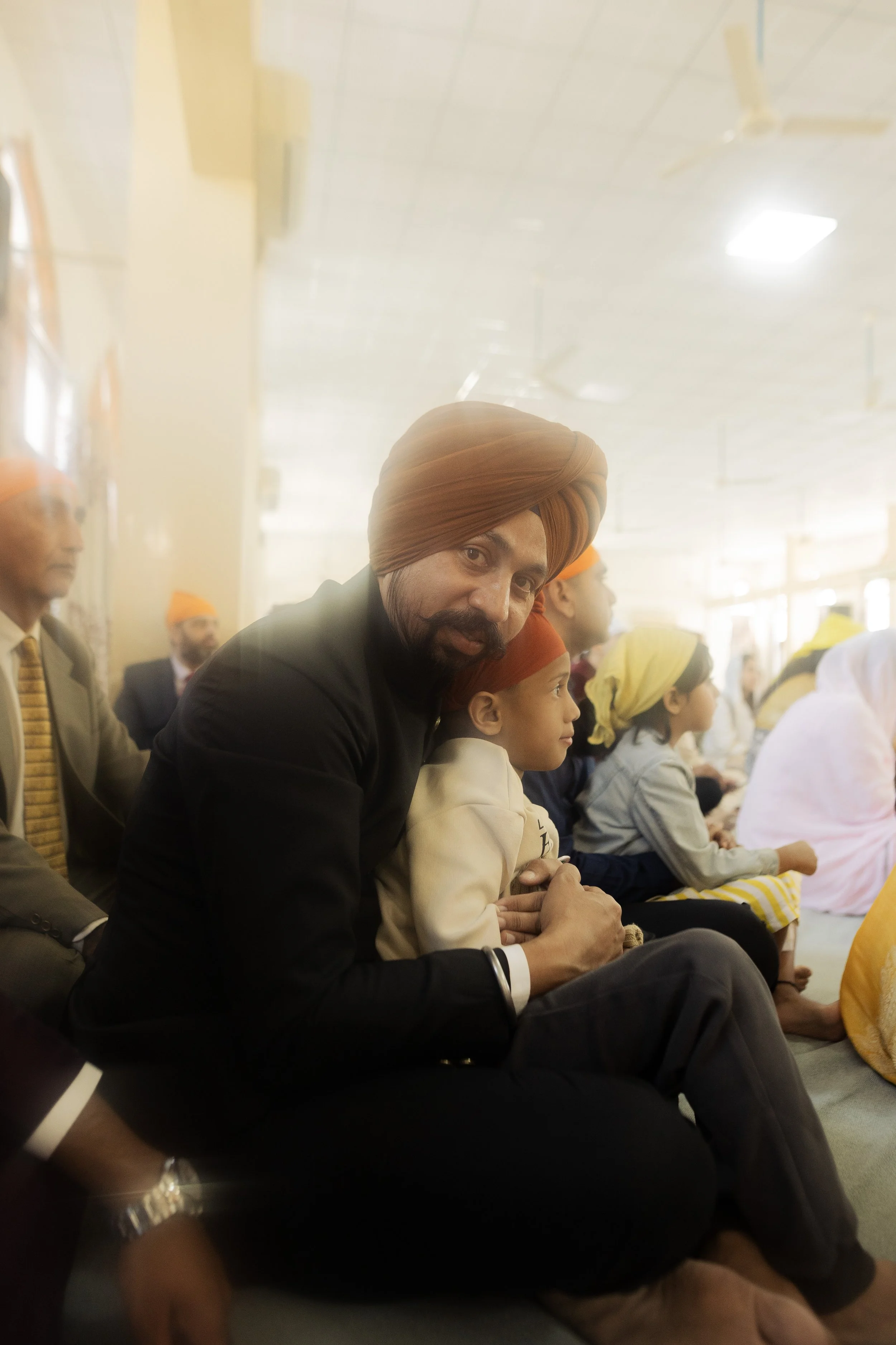 Man wearing a brown turban and black suit sitting on the floor with children around him, in a religious or cultural gathering.