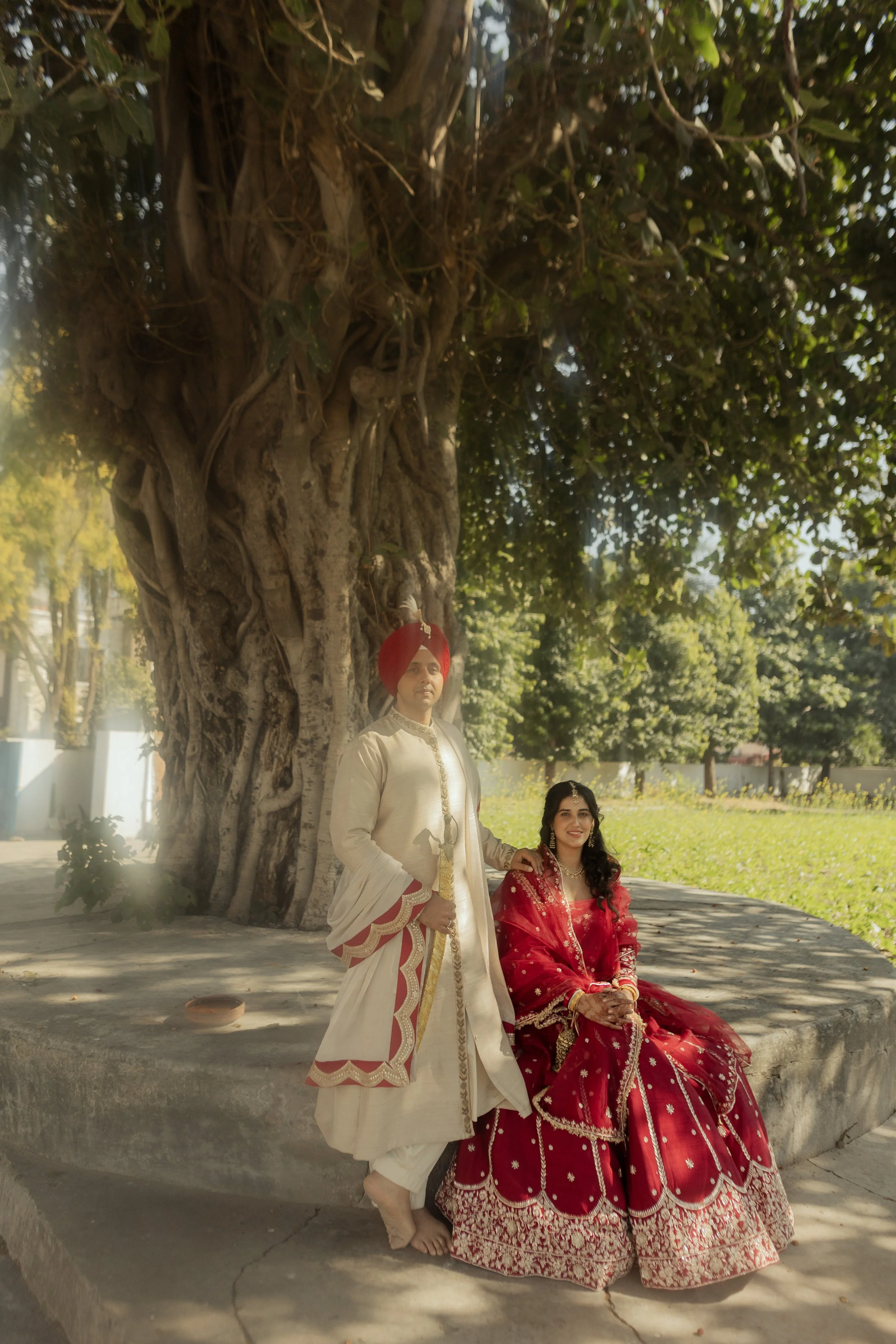 A man dressed in traditional Indian wedding attire with a red turban and a woman in a red bridal lehenga, posing under a large tree in a park.