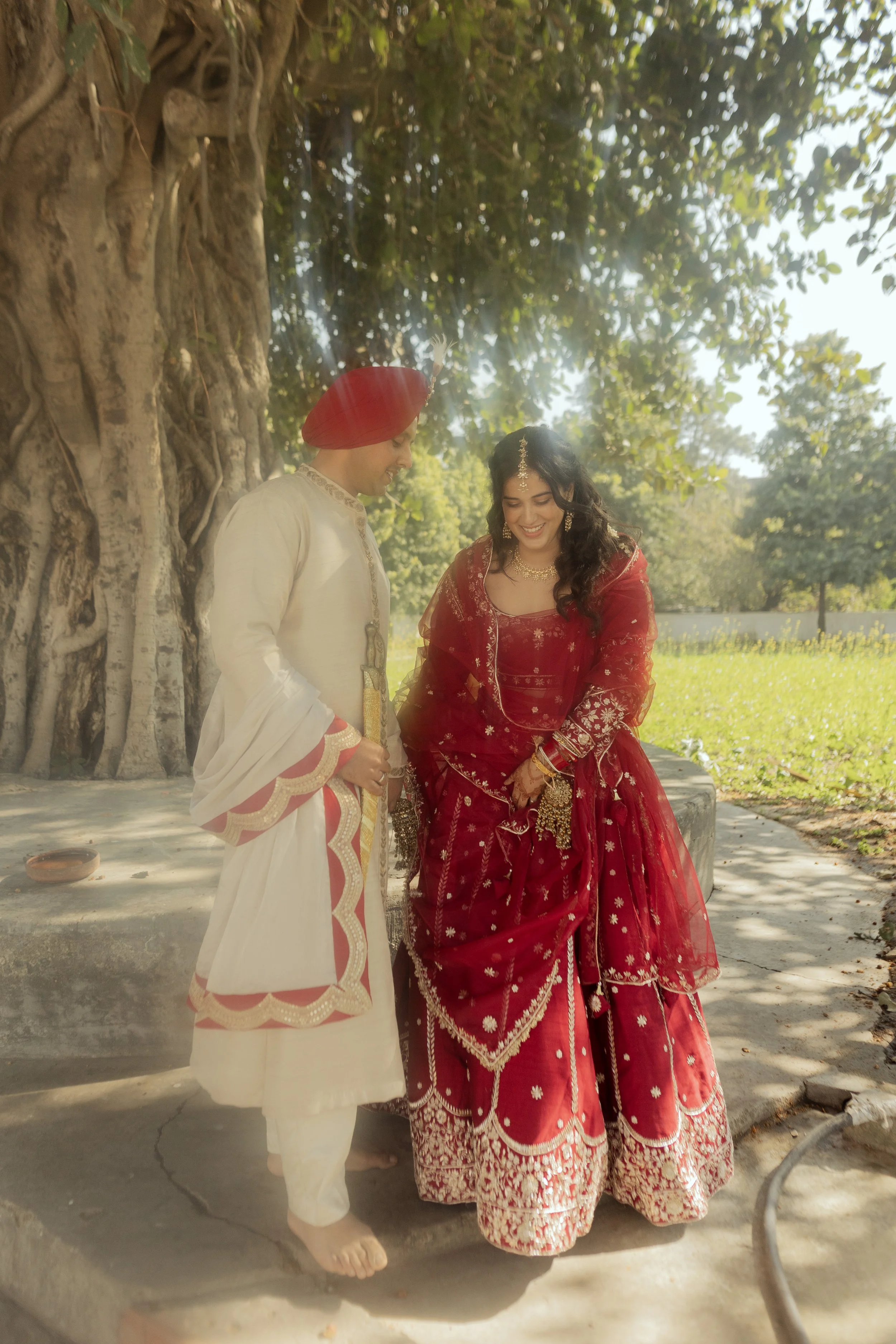 A couple dressed in traditional Indian wedding attire, standing outdoors under a large tree with sunlight filtering through the leaves, smiling and looking at each other.