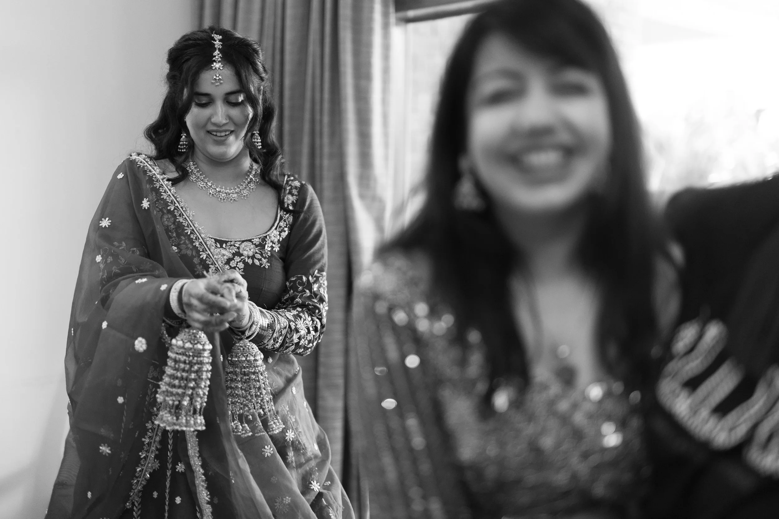 A woman dressed in traditional Indian bridal attire, celebrating and smiling, with another woman taking a selfie, both wearing jewelry and ornate clothing.