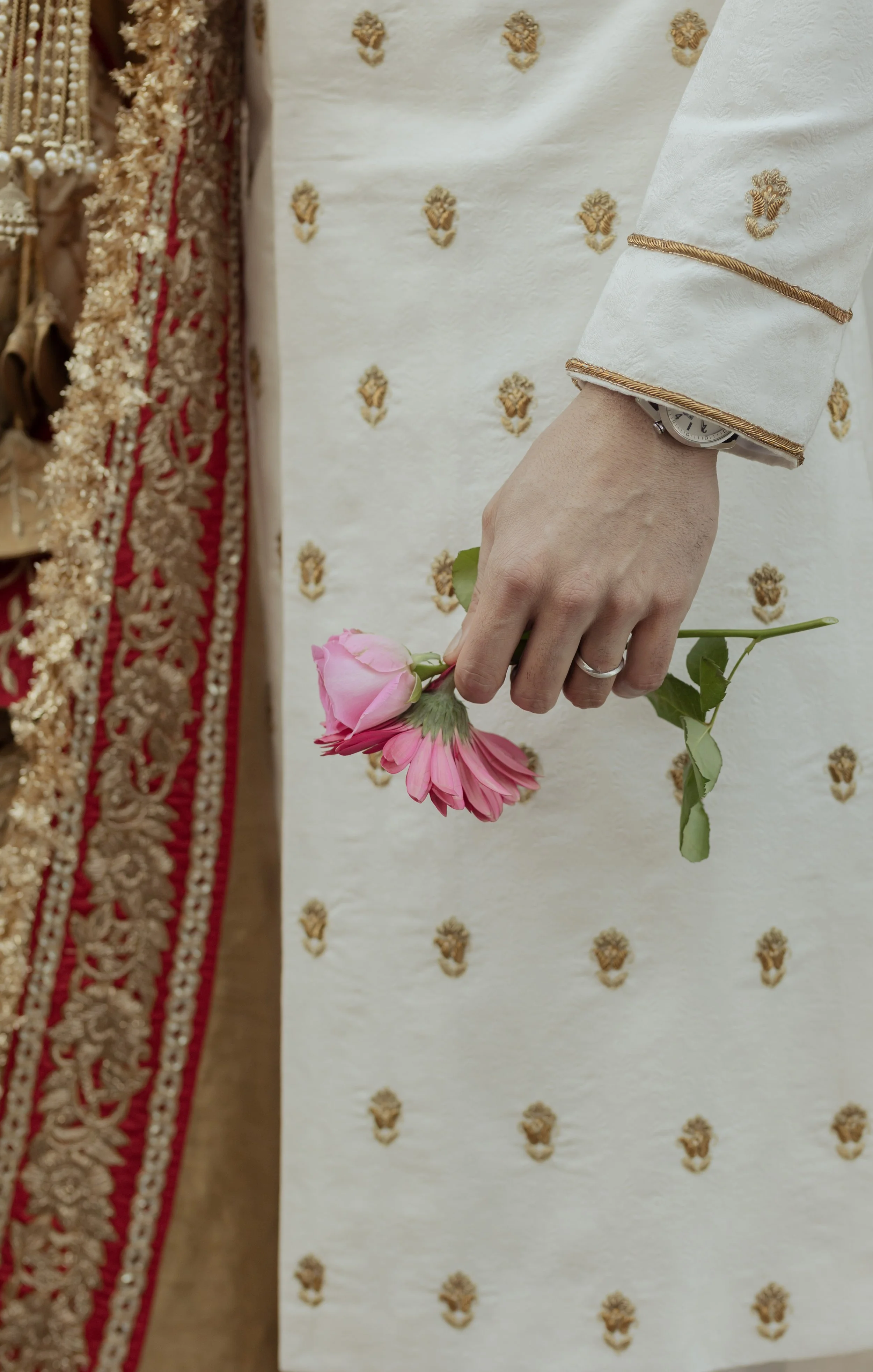 Person holding a pink flower, wearing a white and gold embroidered garment, standing next to another person wearing a gold and red embroidered garment.