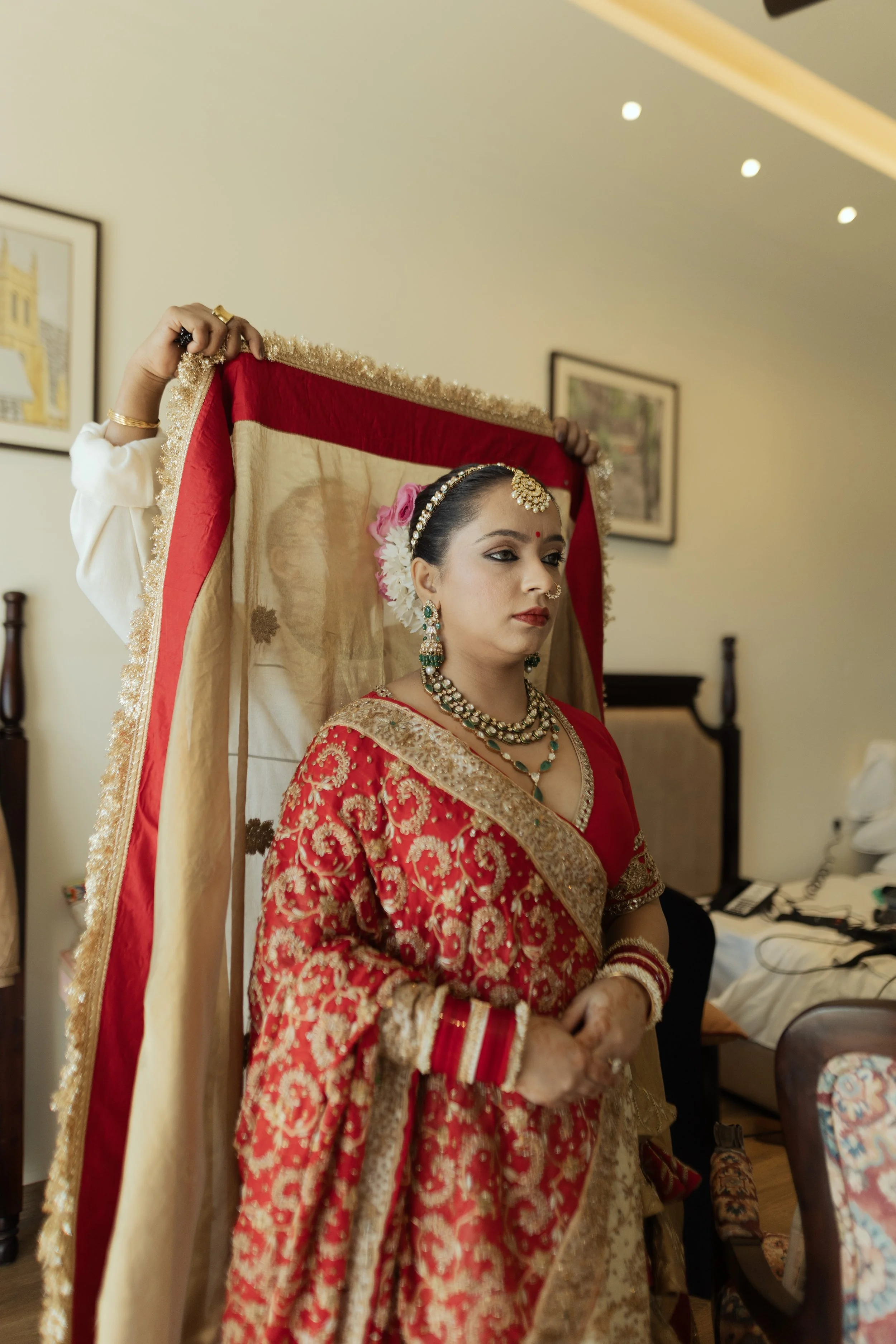 A woman dressed in an ornate red and gold saree with jewelry, sitting with a serious expression, while someone places a decorative backdrop over her head in a room decorated for a celebration.