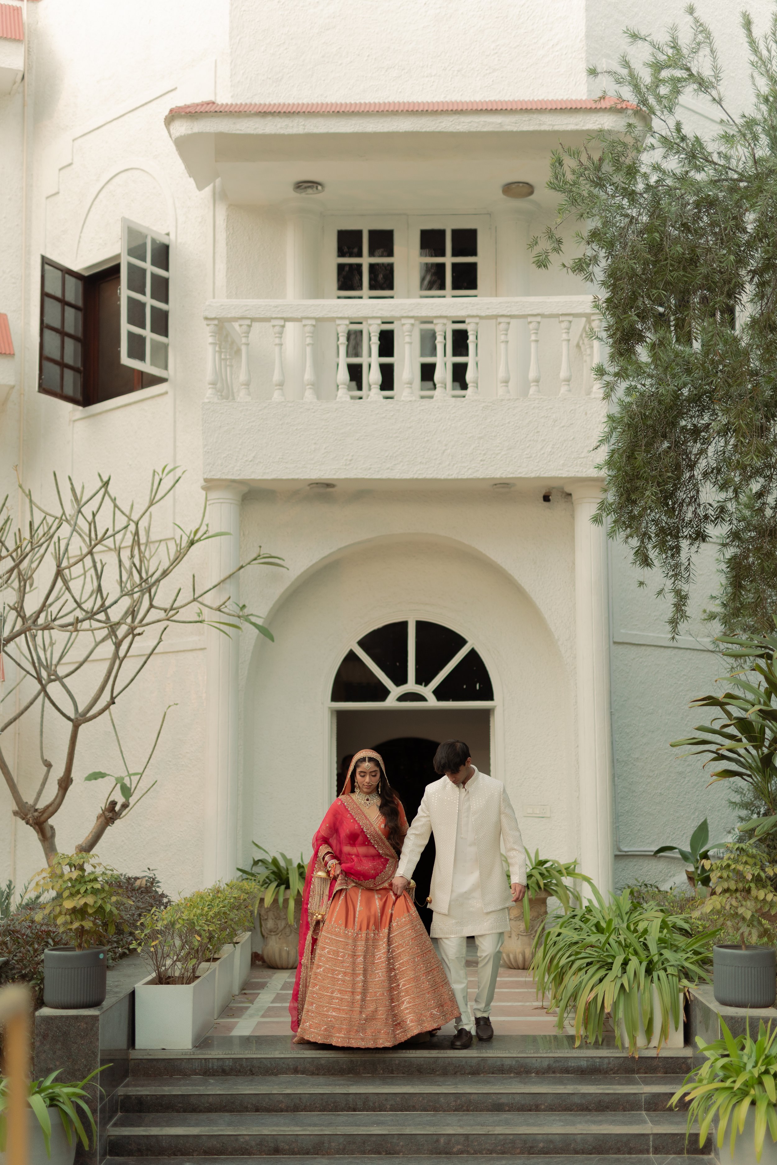 A bride and groom in traditional Indian attire walking down the steps in front of a white house with potted plants and greenery.