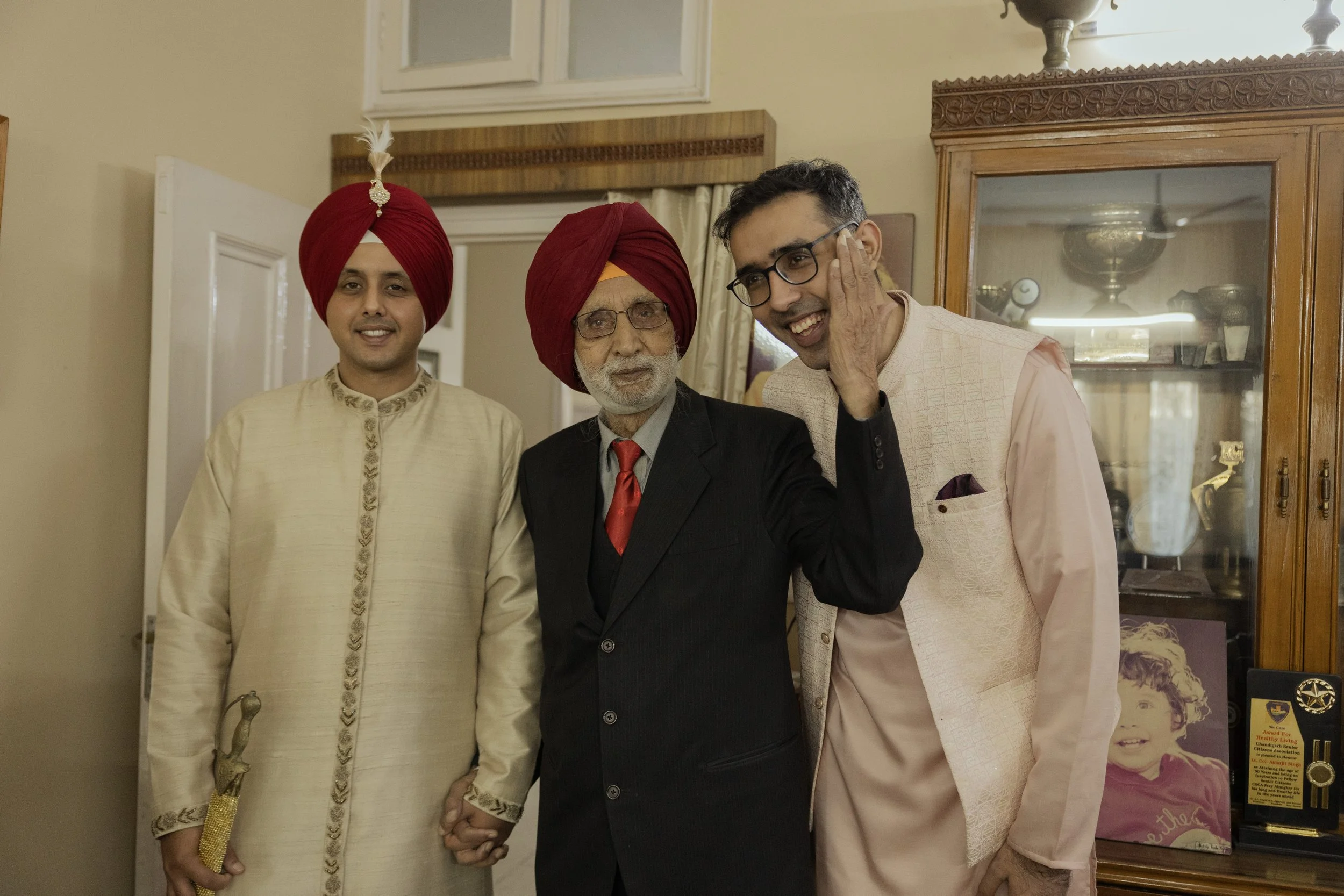 Three men in traditional Indian attire and one man in a suit, standing closely together in a room with wooden furniture and a display cabinet, smiling and celebrating.