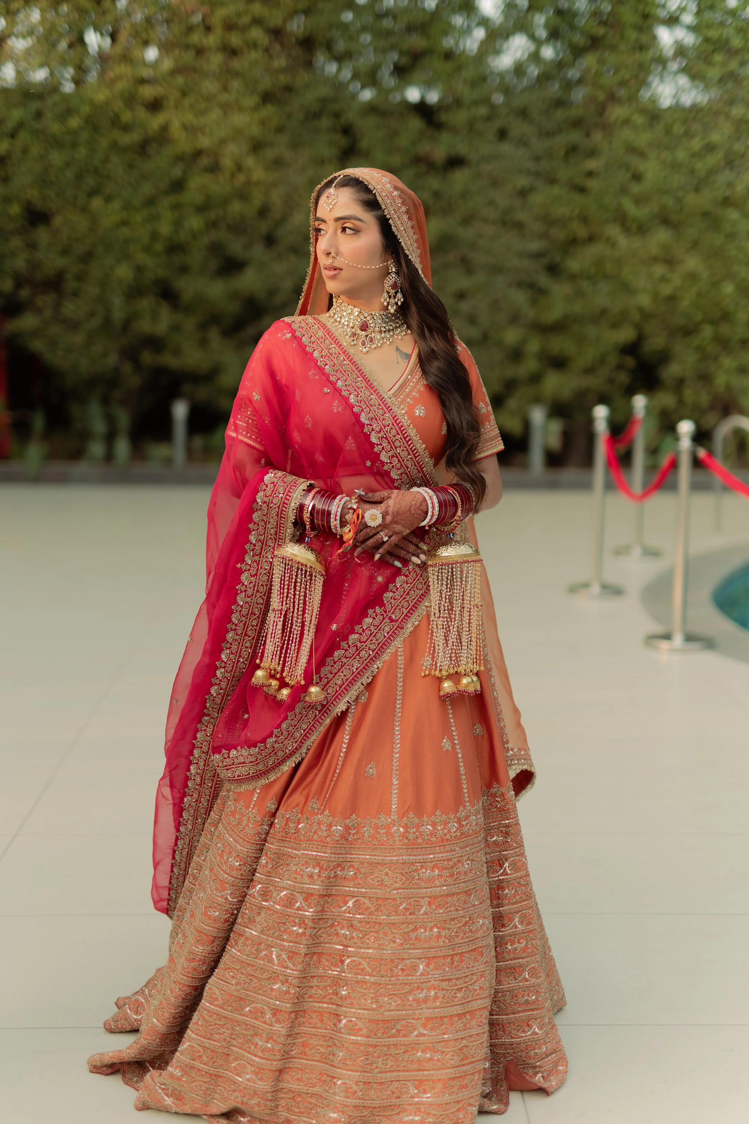 A woman dressed in traditional Indian bridal attire, wearing an orange and red lehenga with gold embroidery, gold jewelry, bangles, and henna on her hands, stands outdoors with trees in the background.