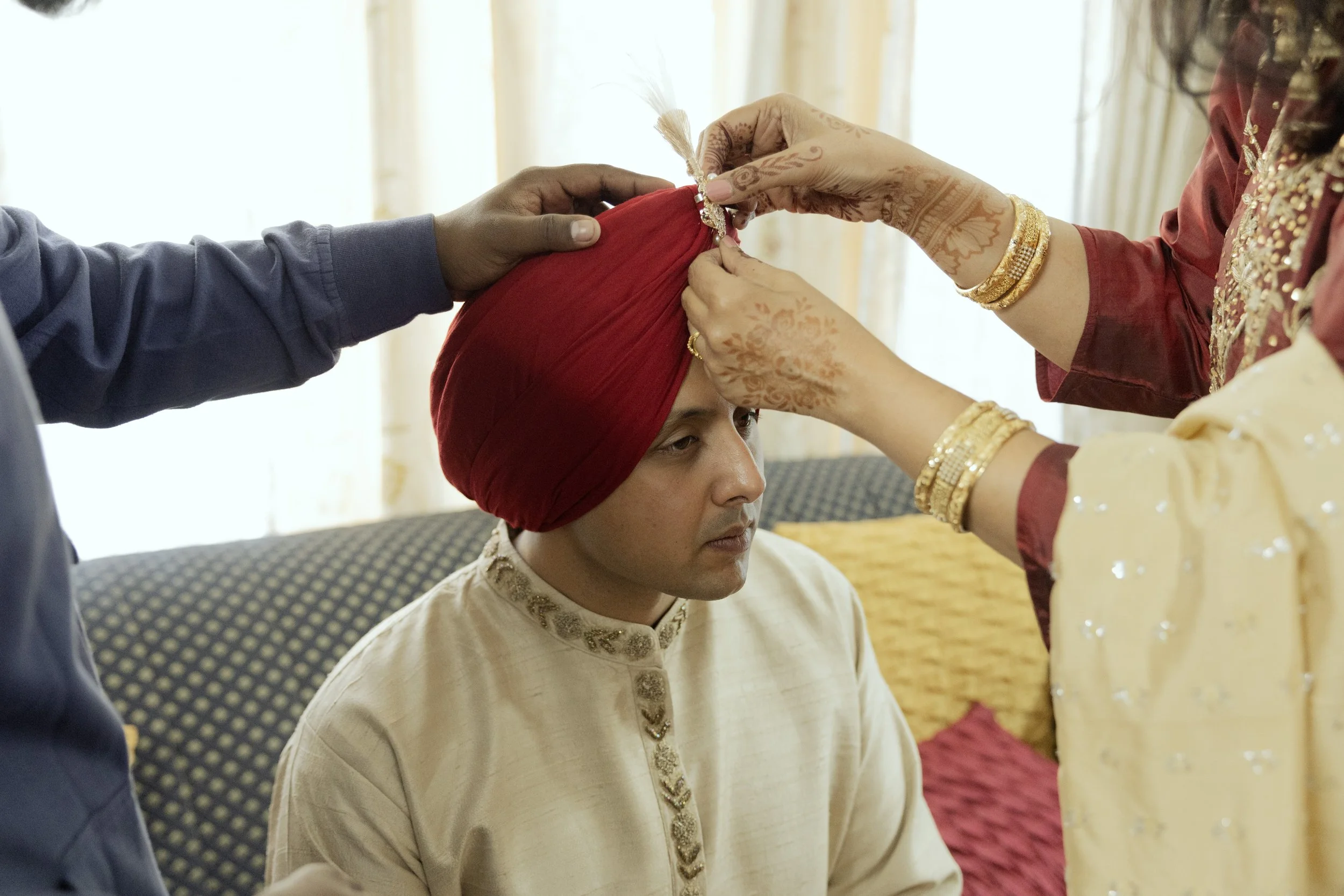 A groom in cream traditional Indian attire and a red turban sits with eyes closed while women, wearing yellow and maroon clothing with gold jewelry, perform a ritual on his forehead.