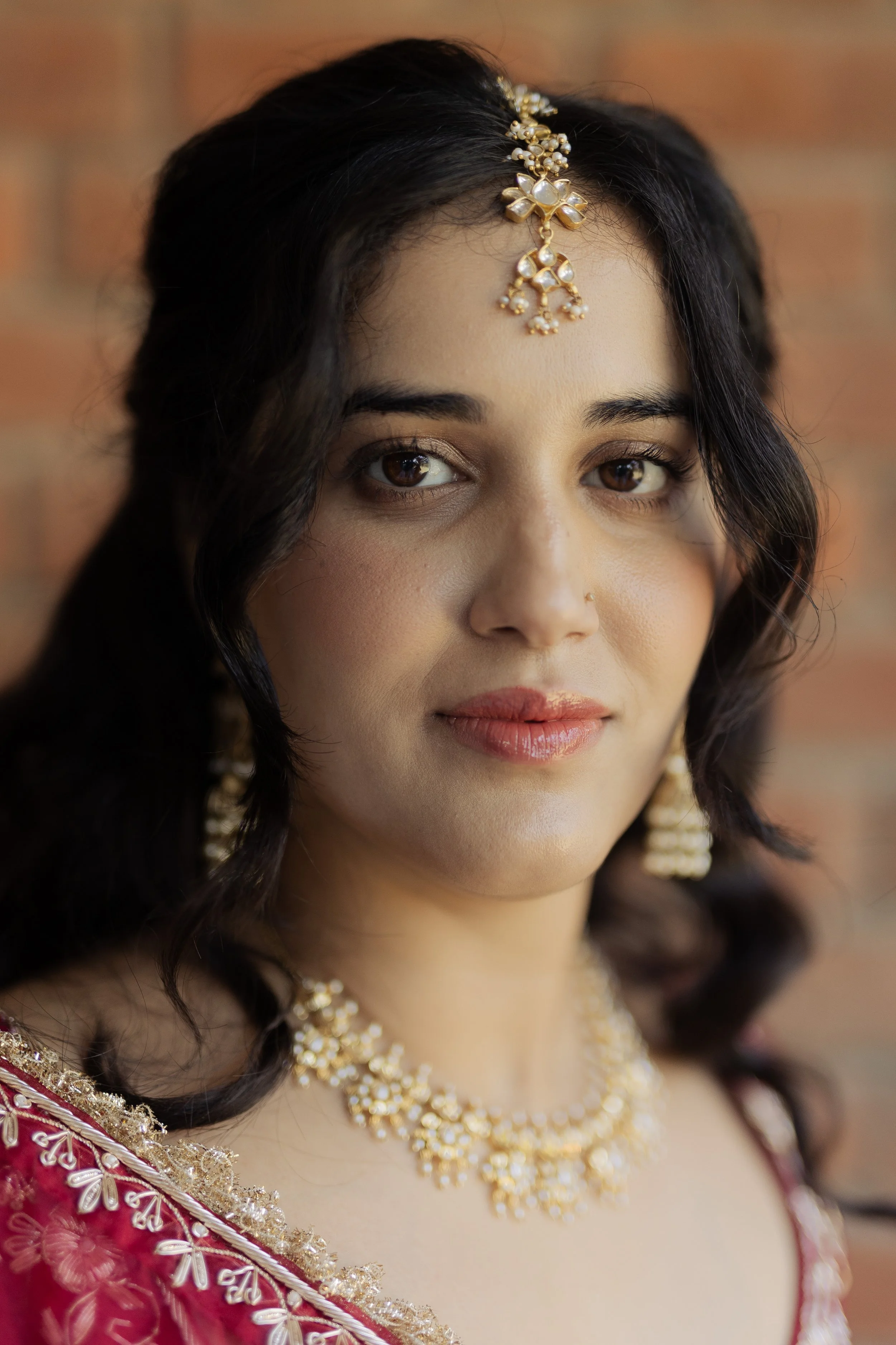 Close-up portrait of an Indian woman wearing traditional gold jewelry and a red embroidered saree, standing against a brick wall.