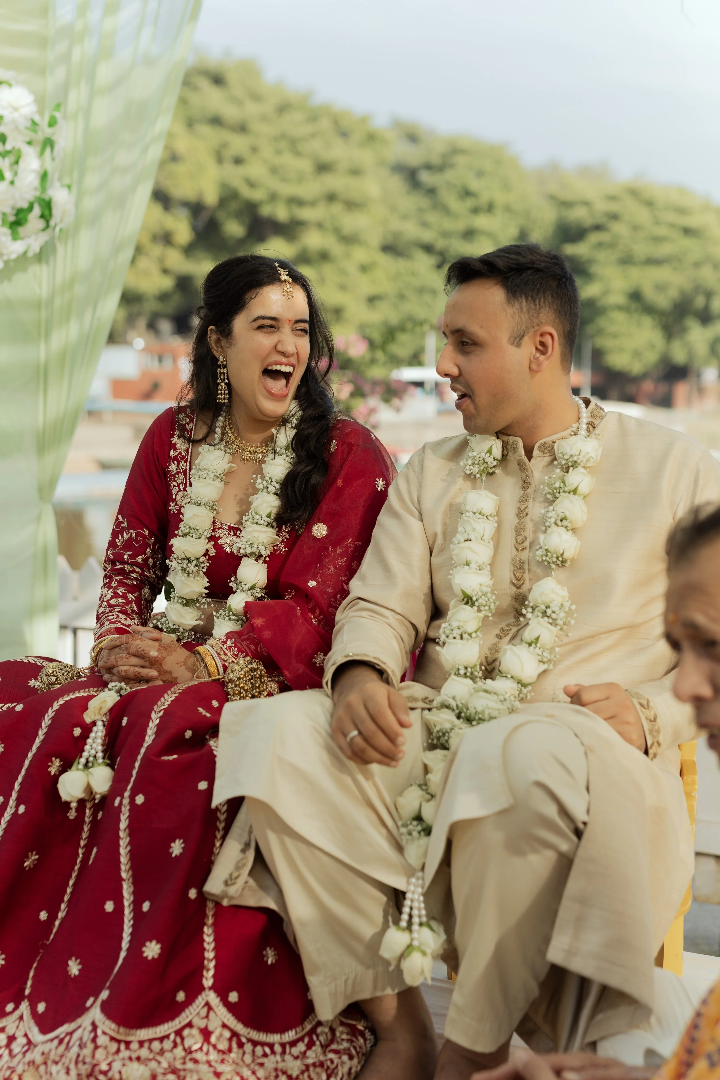 A happy bride and groom sitting together during an outdoor wedding ceremony, dressed in traditional Indian attire with floral garlands.