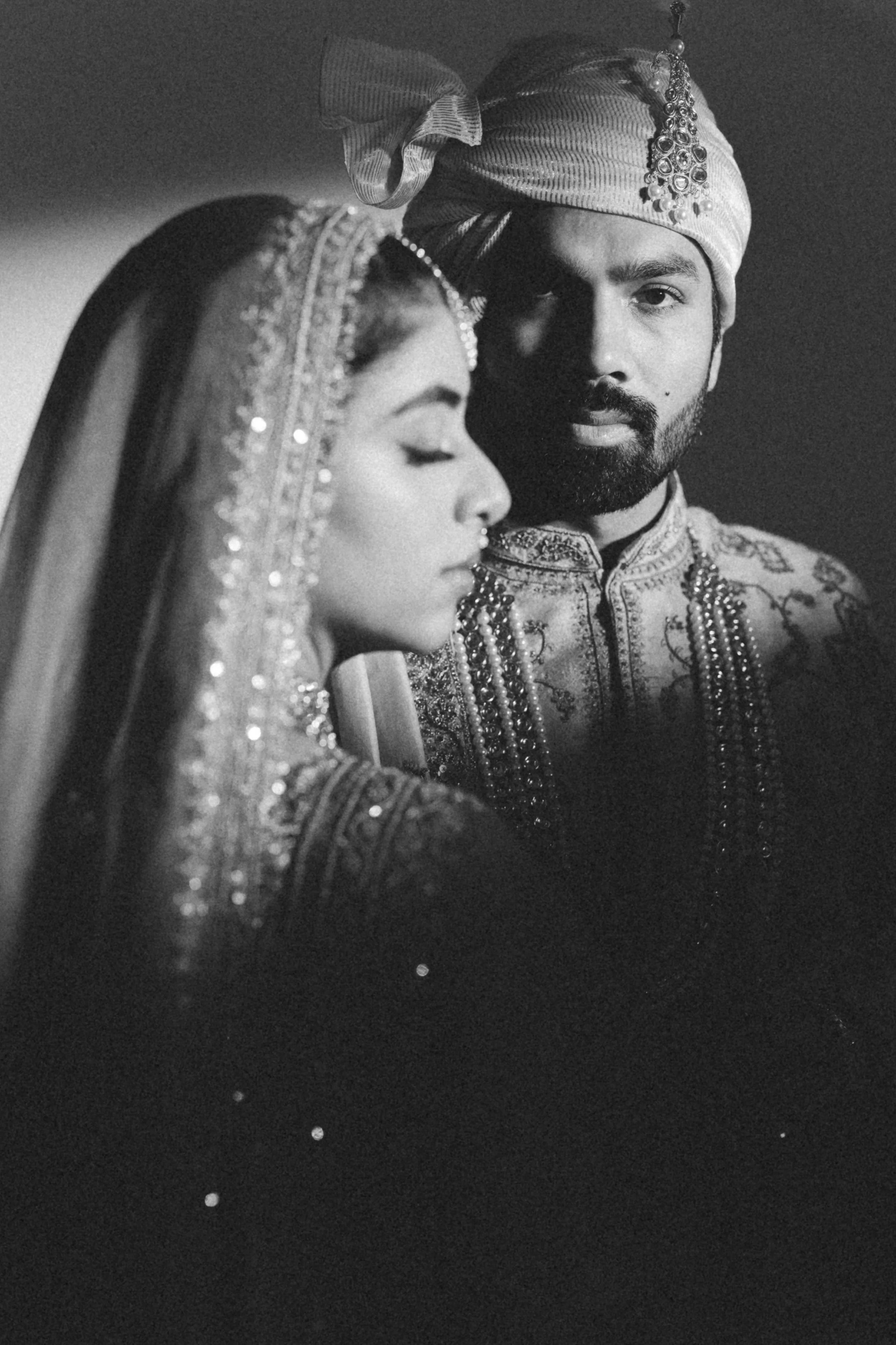A black and white photo of a bride and groom in traditional Indian wedding attire. The bride has her eyes closed and is wearing a decorated veil, jewelry, and a forehead ornament. The groom has a beard, is looking directly at the camera, and is weari