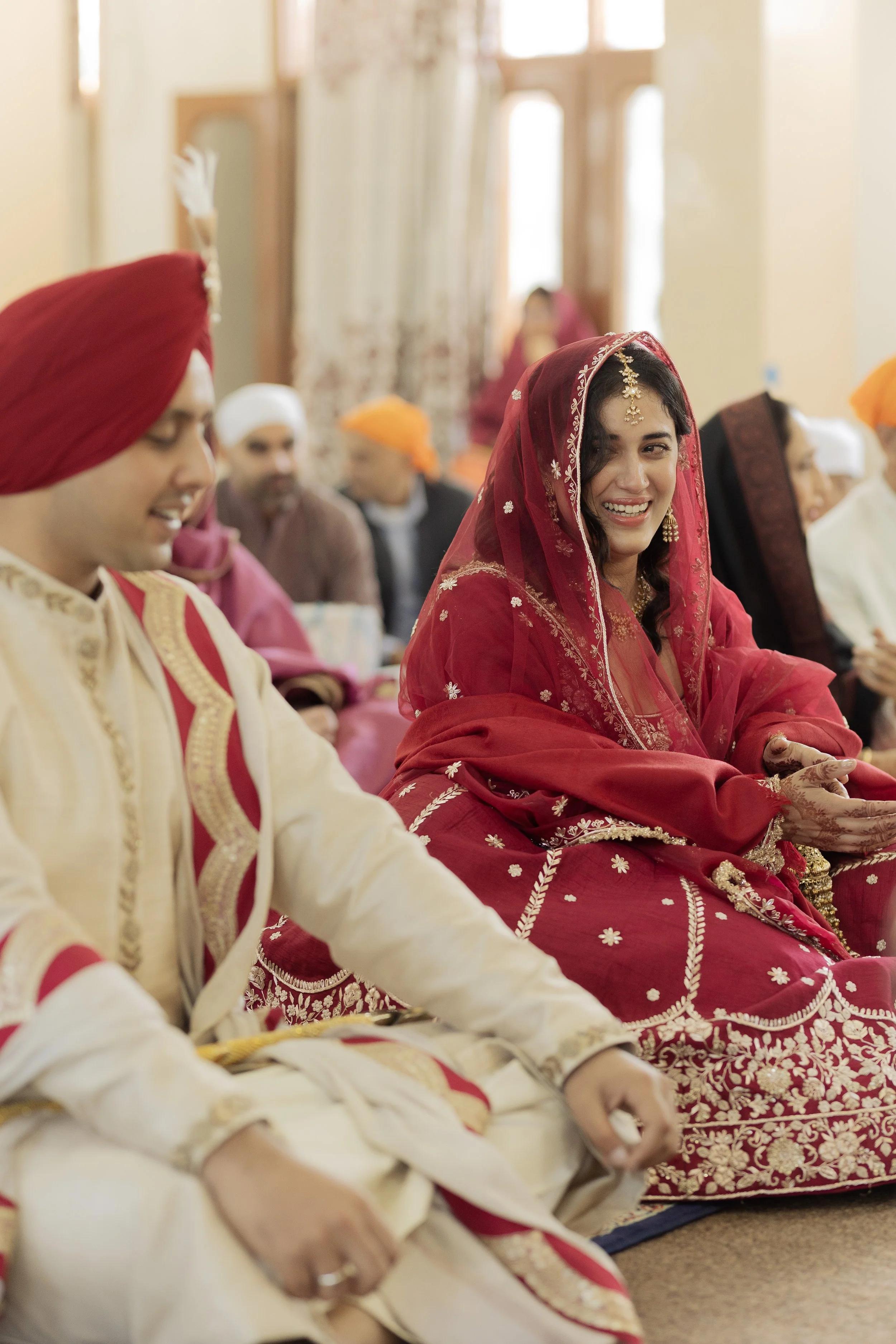 Indian wedding ceremony with a woman in a red embroidered saree and veil smiling, sitting next to a man in traditional cream and red attire, surrounded by other guests in colorful traditional clothing.