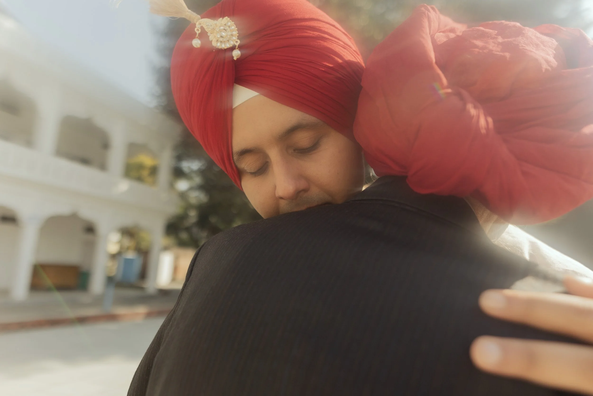 A person wearing a red turban embracing someone with their eyes closed outdoors.