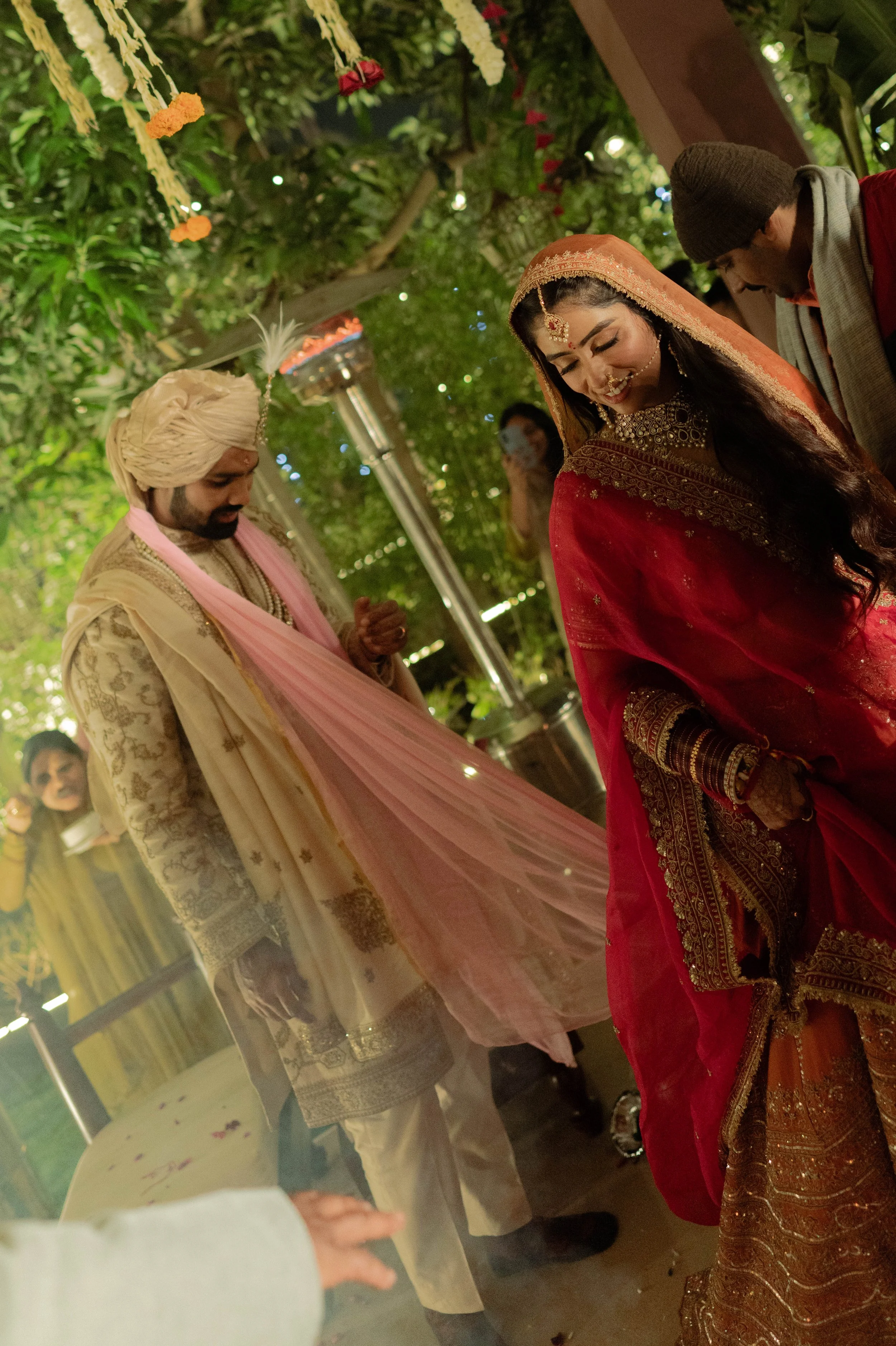 Indian bride in a red saree smiling during a traditional wedding ceremony, with groom dressed in cream and pink attire, under decorated green foliage.