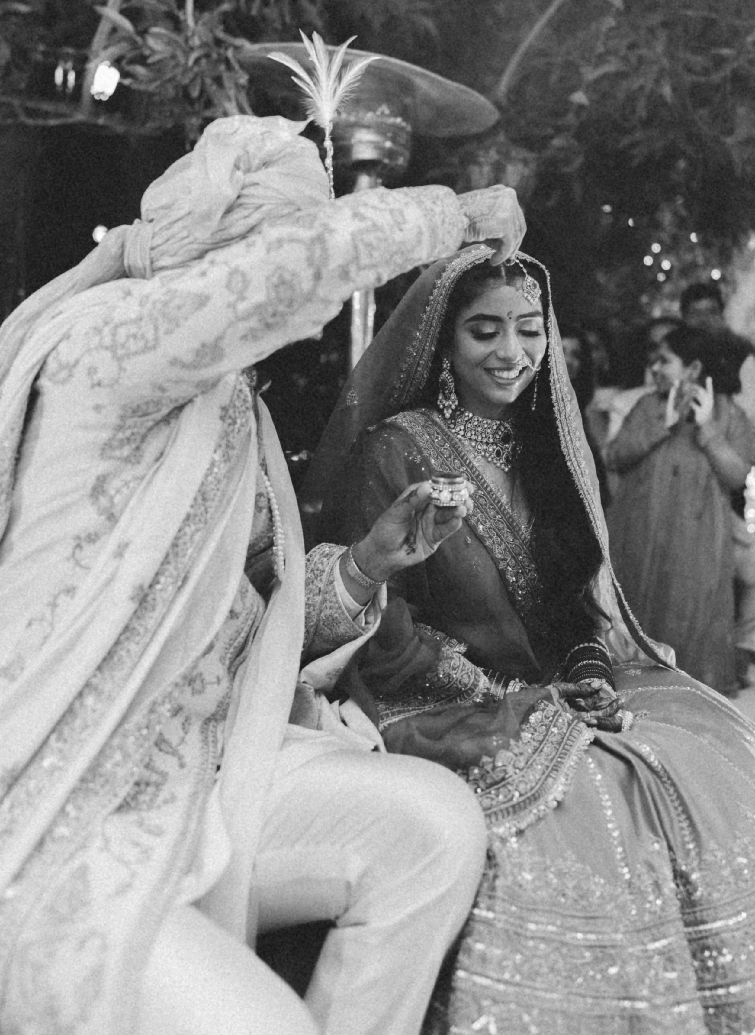 A woman in traditional Indian wedding attire smiling at a wedding ceremony, with a person in traditional clothing tossing flower petals over her head.