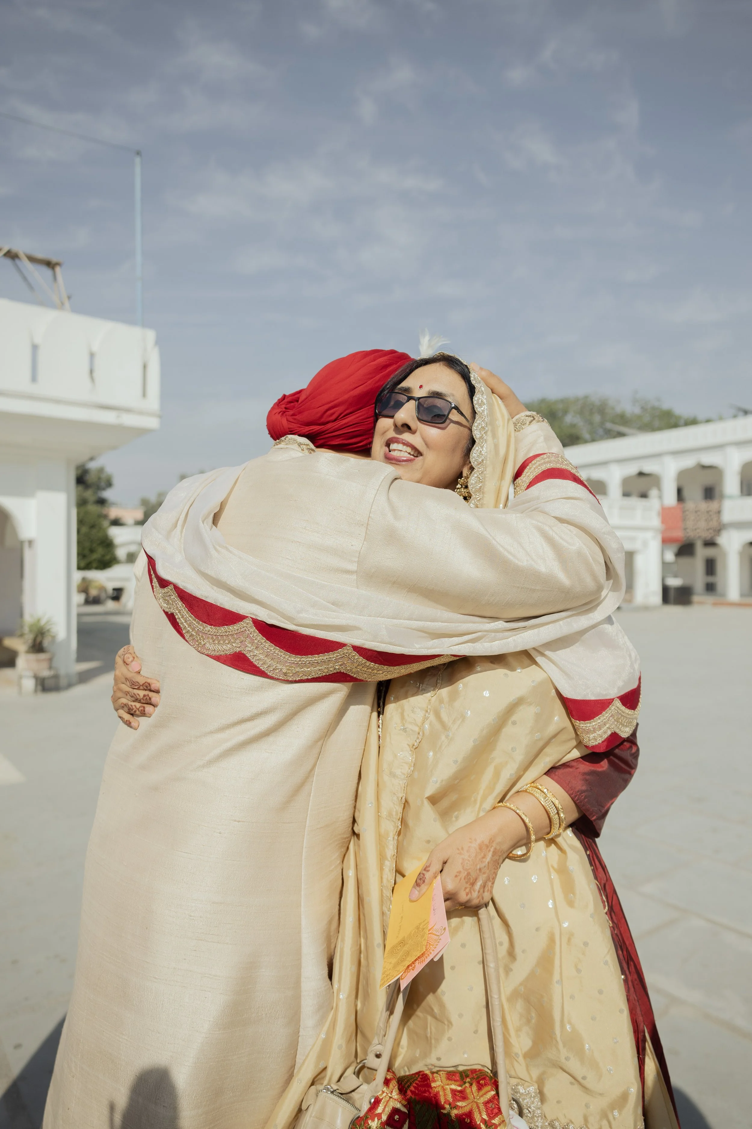 Two women hugging, dressed in traditional Indian attire, outdoors with a clear sky and white buildings in the background.