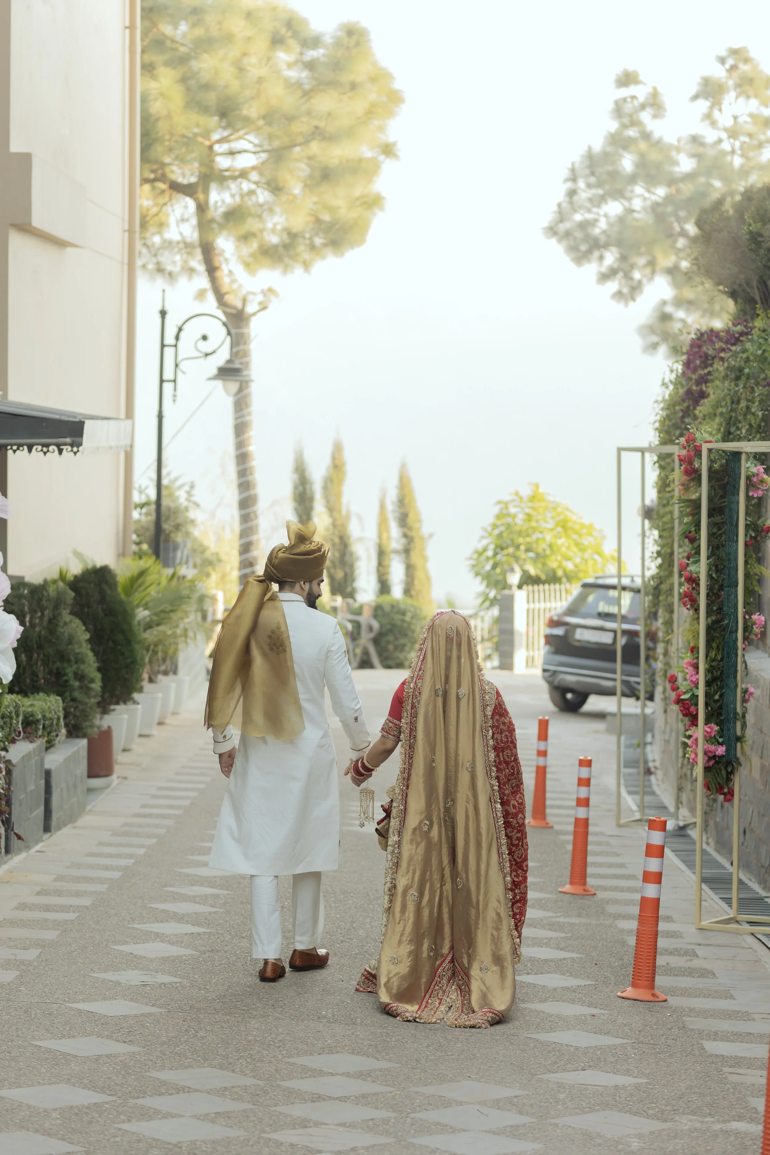 A newly married couple in traditional Indian wedding attire walking hand in hand in an outdoor setting with greenery and plants.