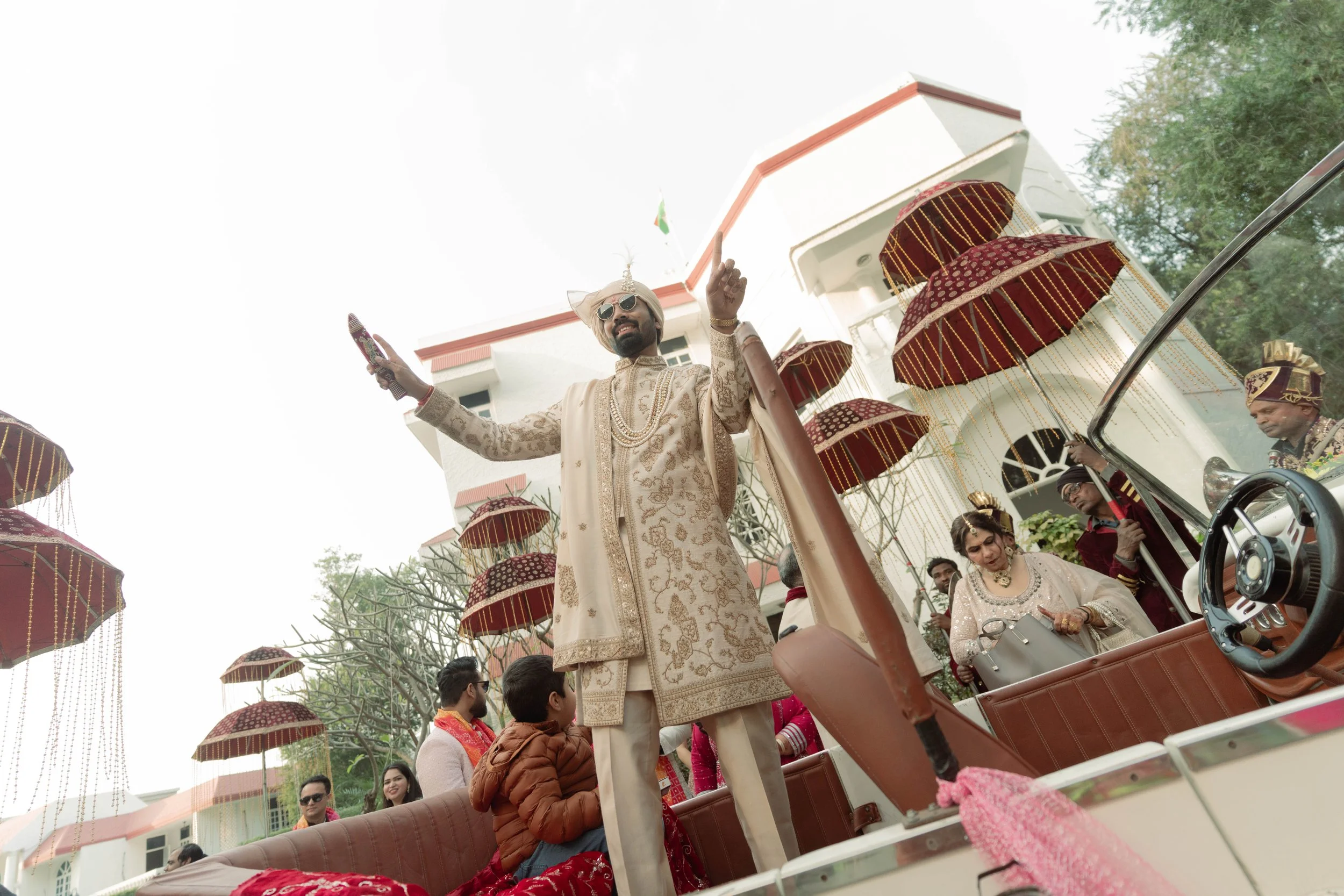 Indian groom in traditional attire standing on a decorated vehicle surrounded by family and friends during a wedding celebration with umbrellas.