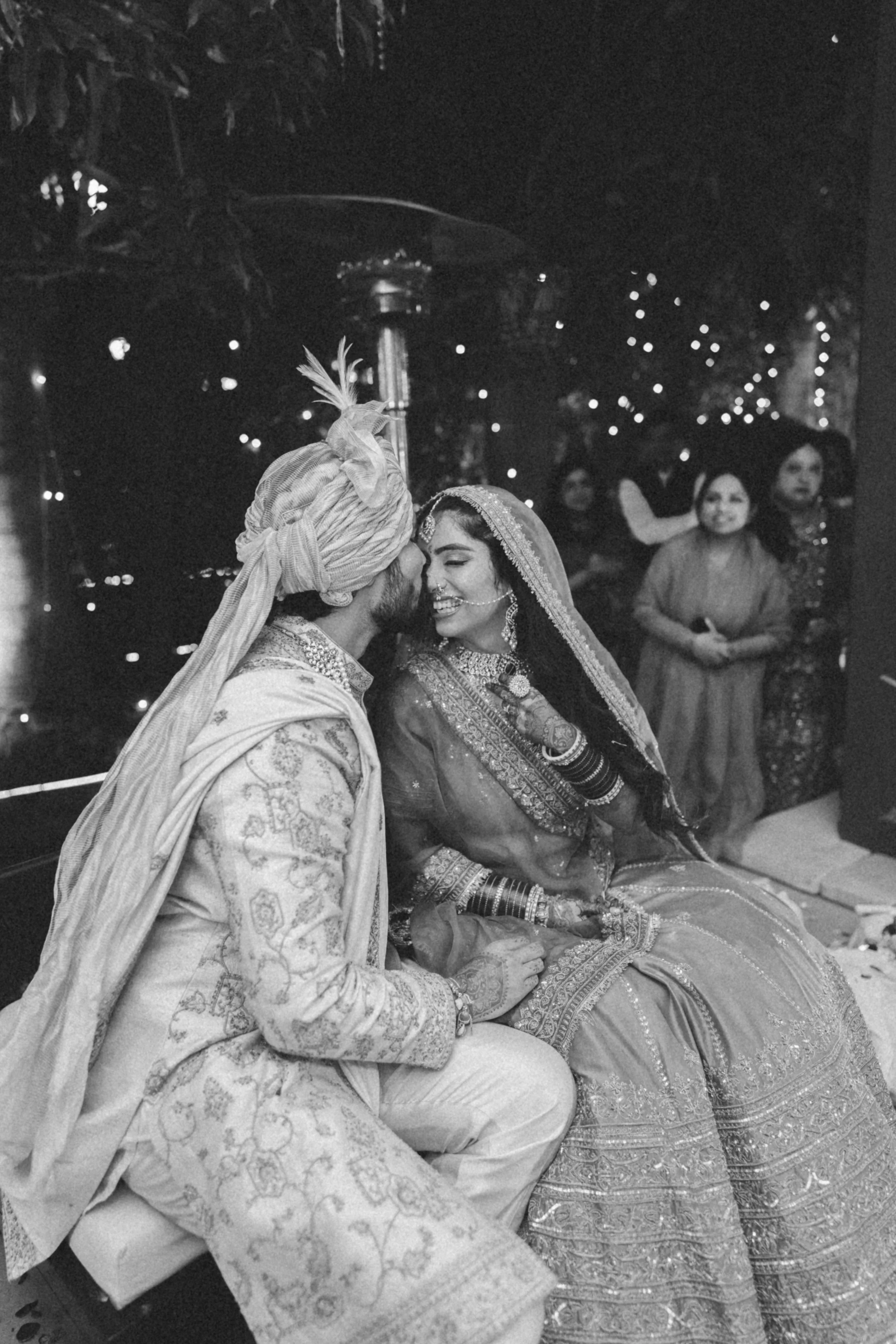A black and white photo of a couple in traditional South Asian wedding attire sharing a kiss at their wedding ceremony, with several women in the background smiling and observing the moment.