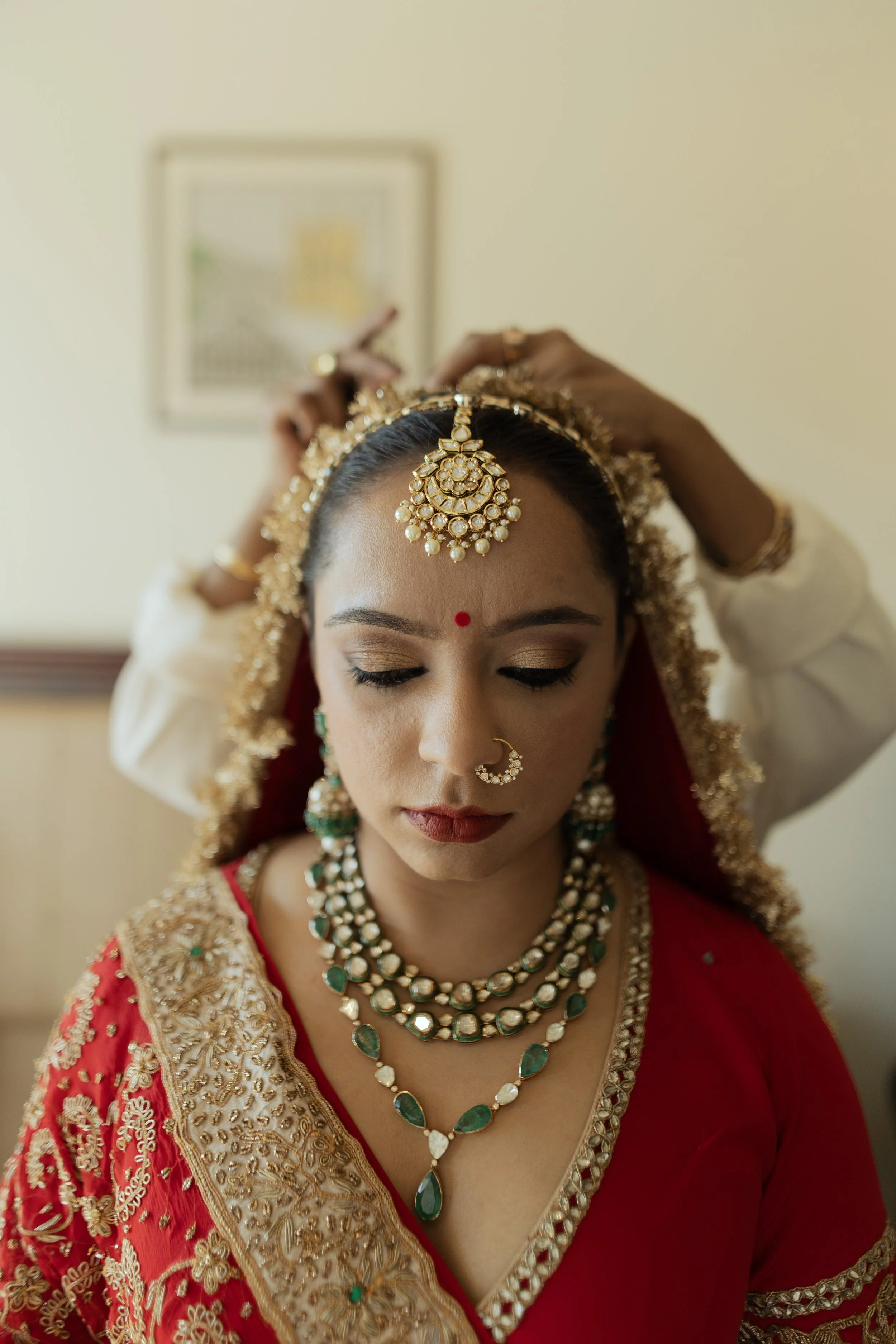 A bride in traditional Indian attire with jewelry and a red saree having gold embroidery, being assisted with her headgear.
