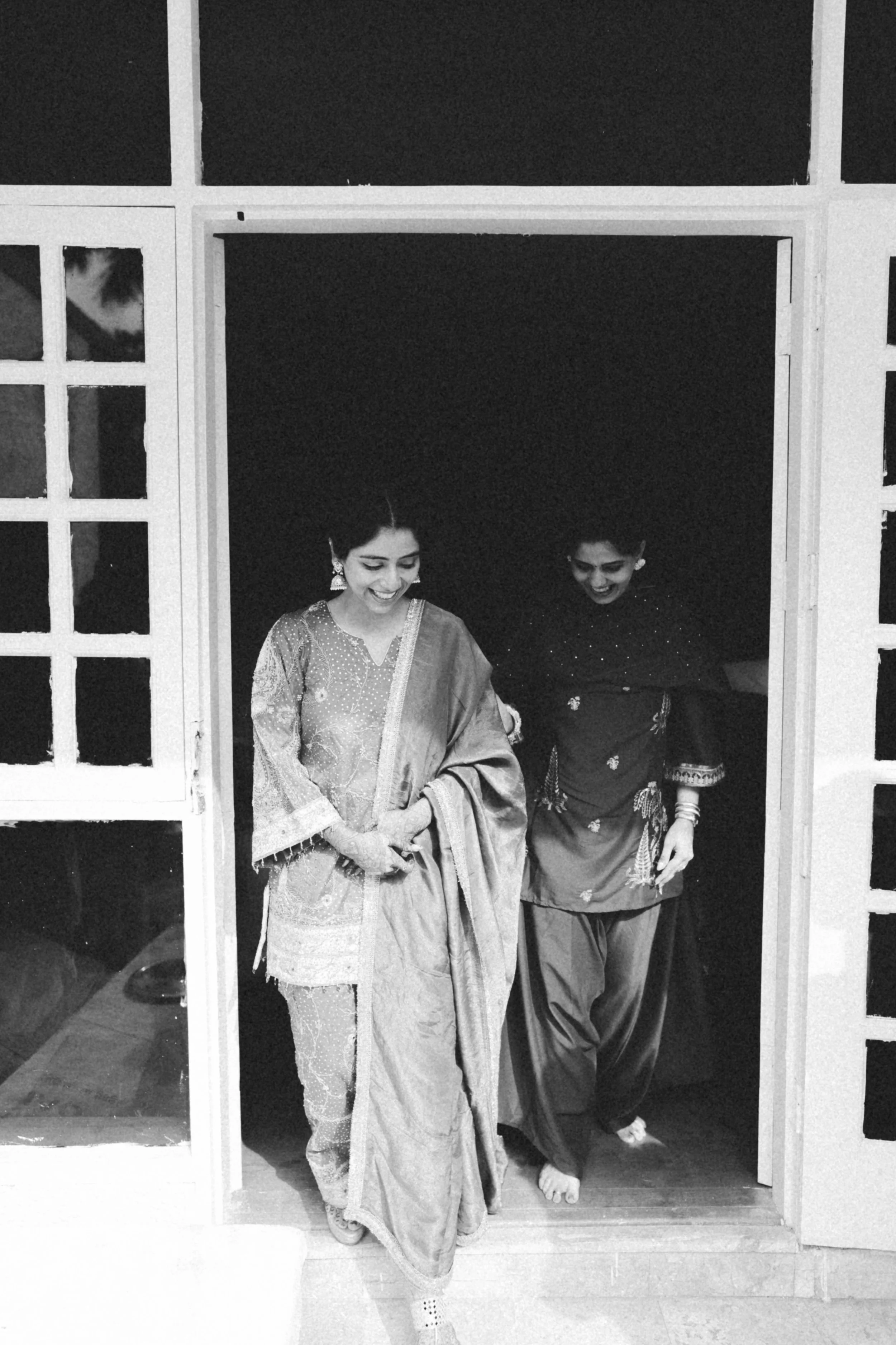 Two women in traditional South Asian attire smiling as they walk out of a doorway.