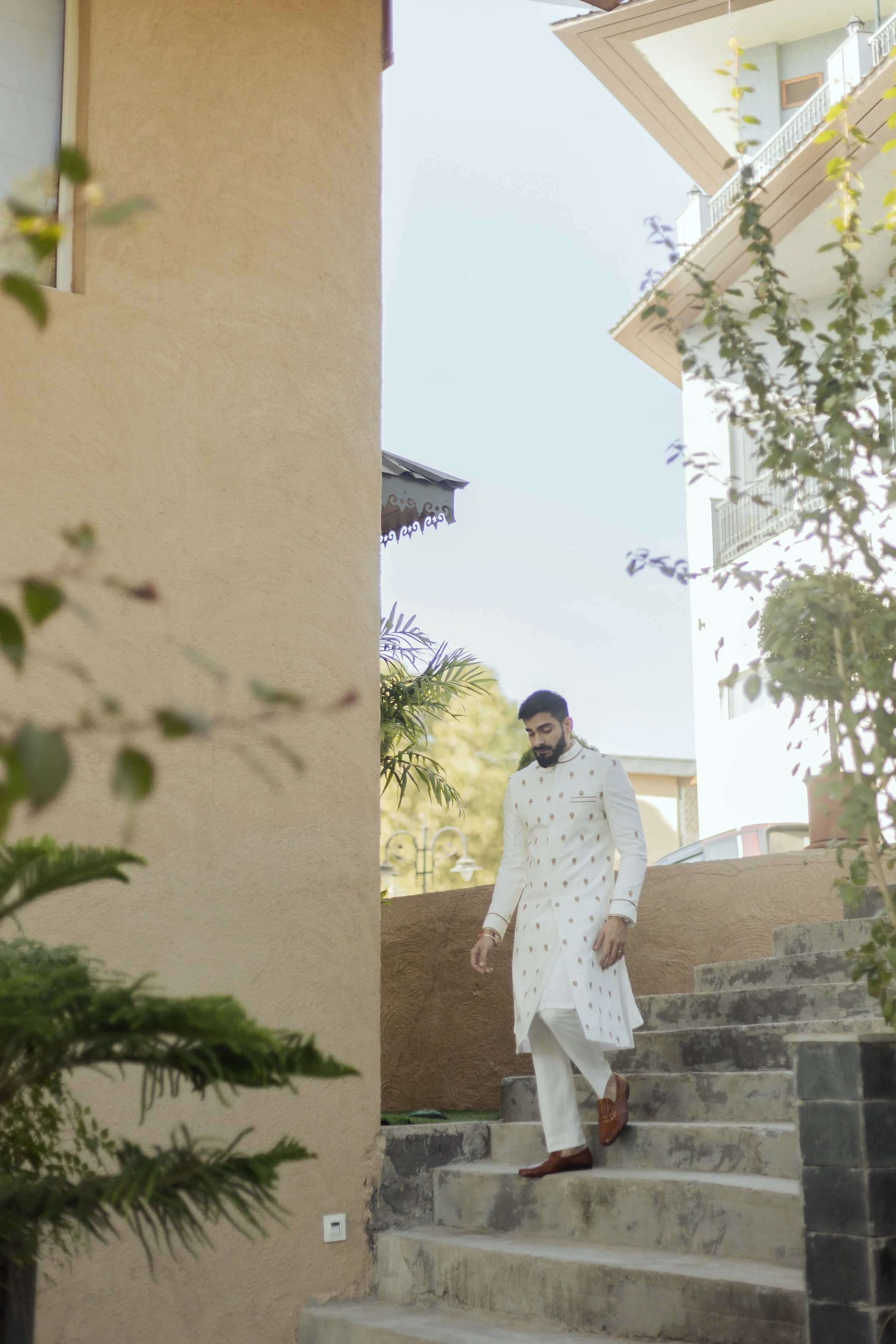 A man wearing traditional white Indian attire with gold accents, descending outdoor stone steps amid greenery with beige walls and neighboring buildings in the background.