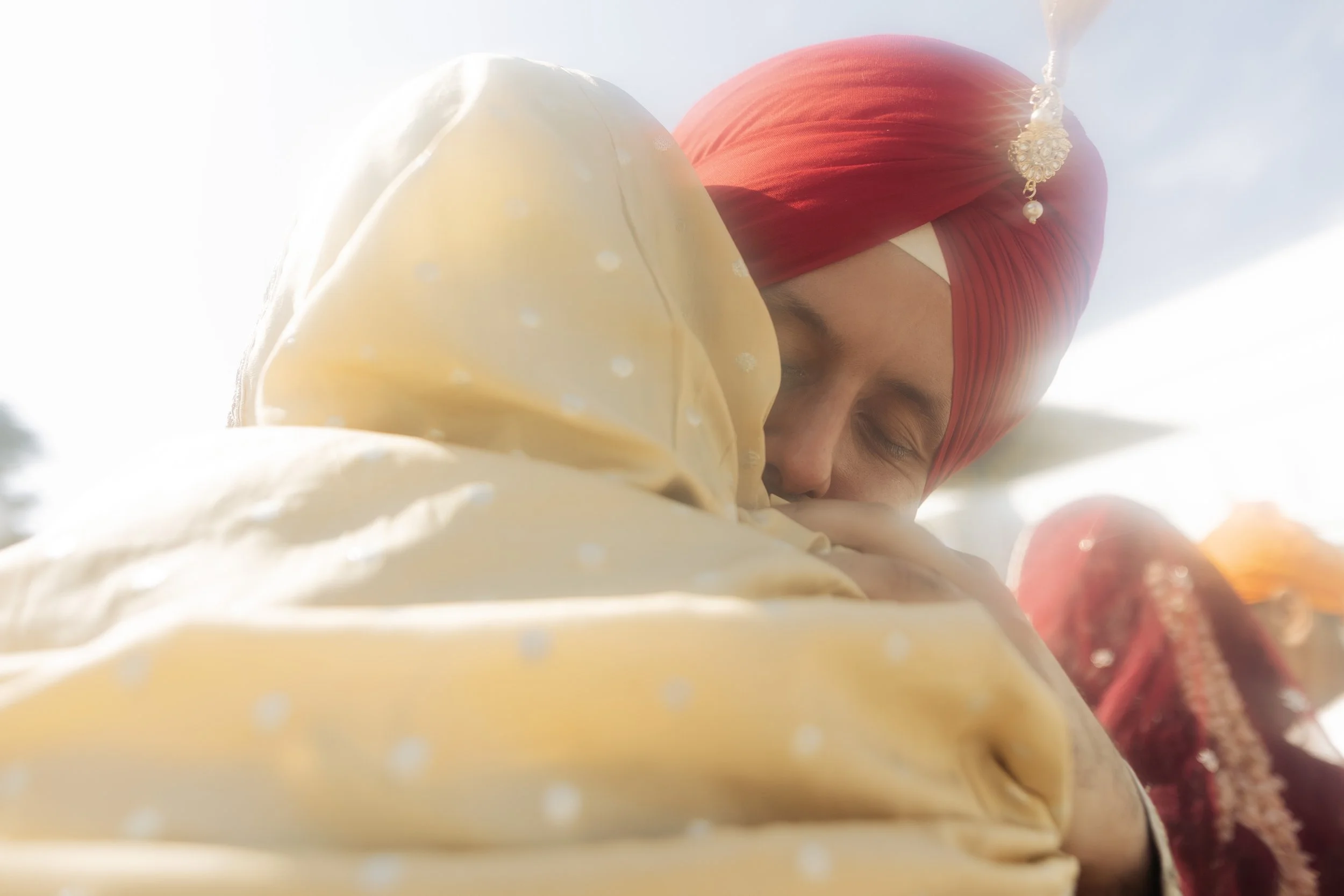 A person dressed in traditional Indian wedding attire with red turban embracing another individual. The scene is bright with sunlight.