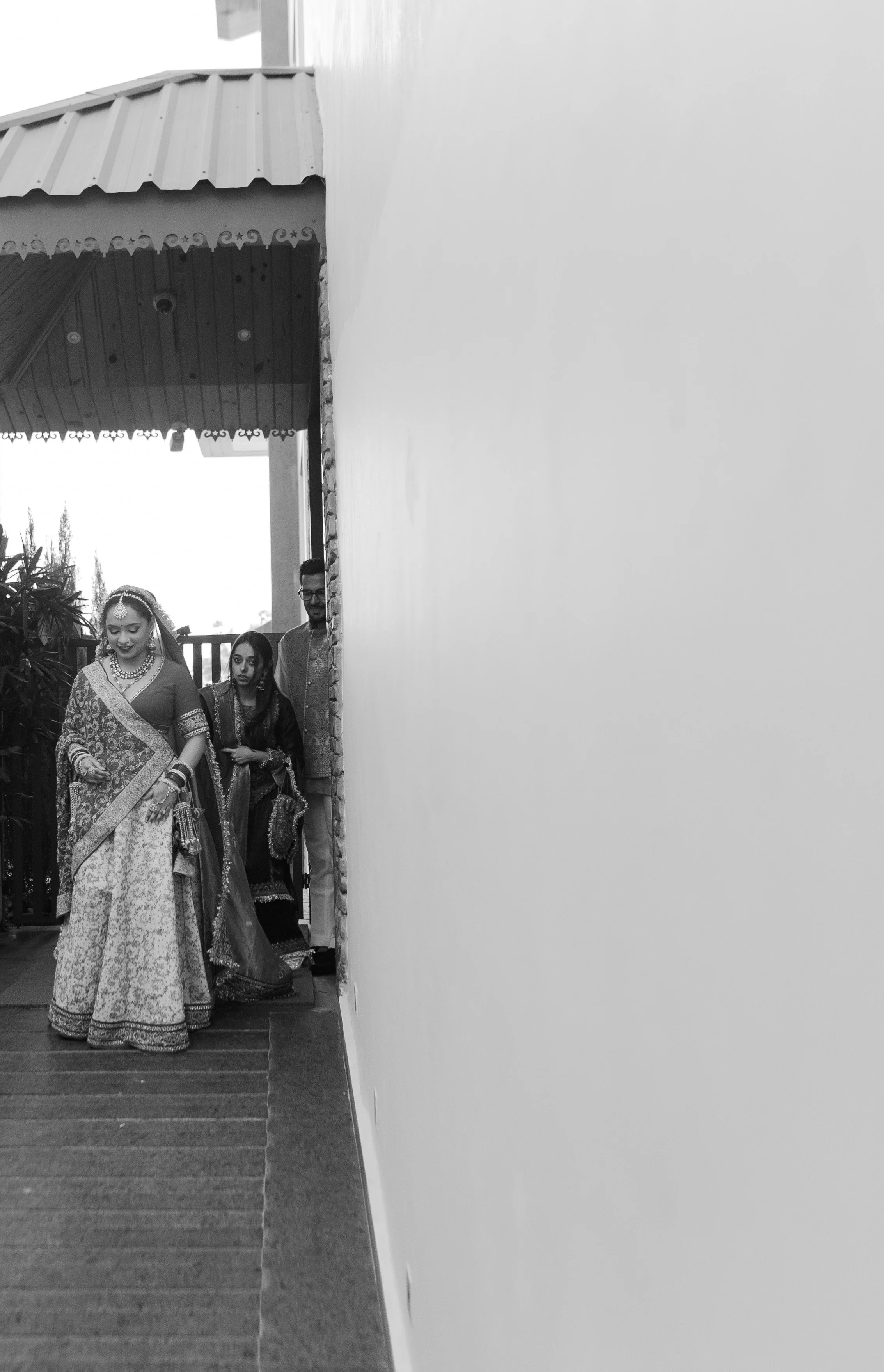 Three people, two women in traditional attire and a man in a suit, walking on a porch with a wooden railing and some plants in the background.